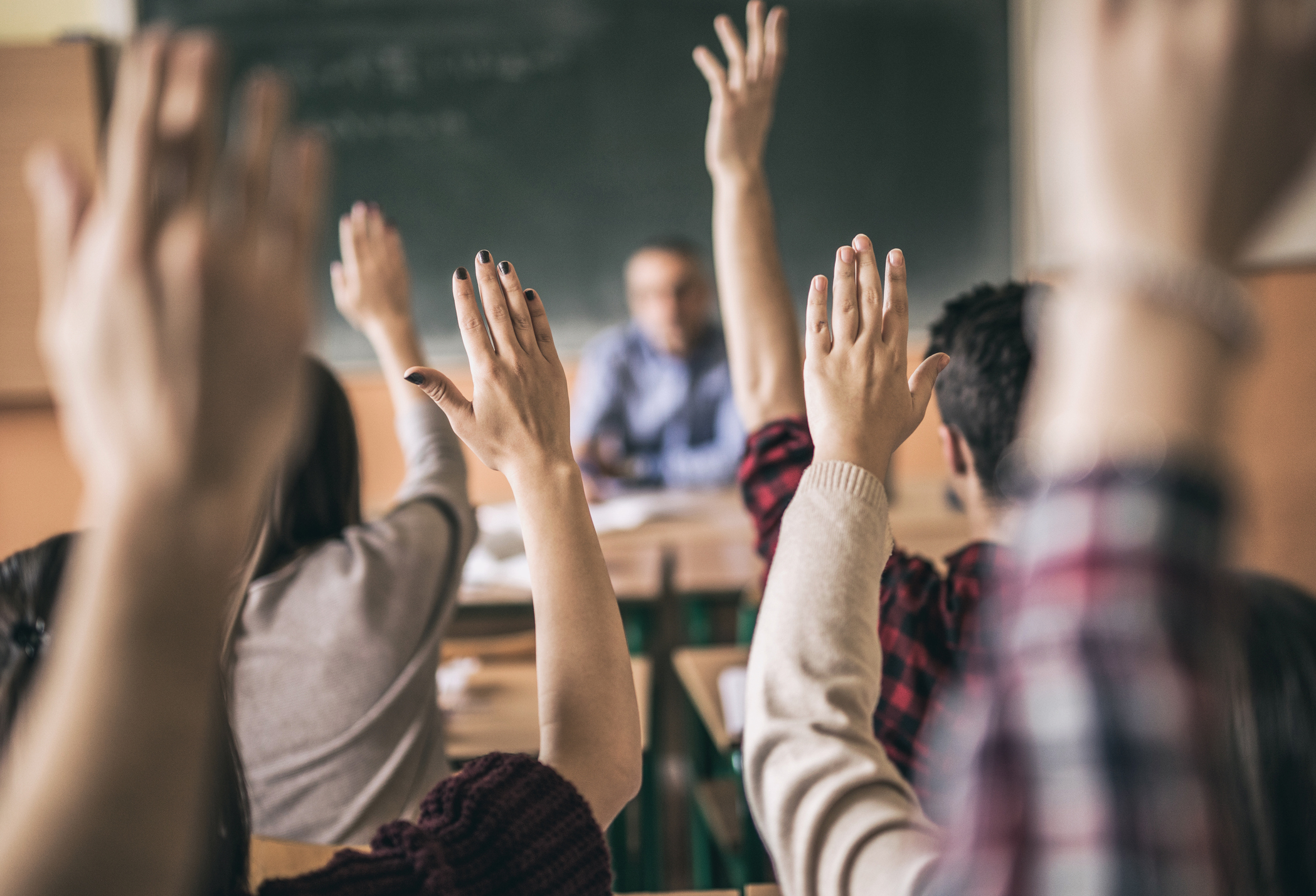 Students in a classroom raising their hands to participate, with a teacher sitting at the front