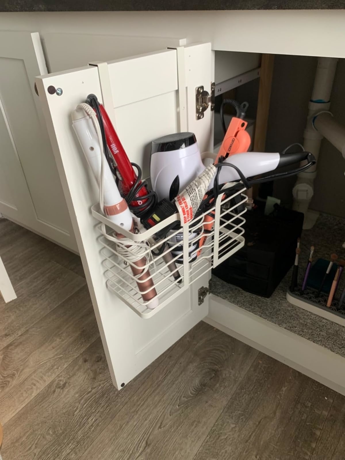 White wire basket inside a bathroom cabinet door holding hair styling tools like a straightener, curling iron, and hair dryer