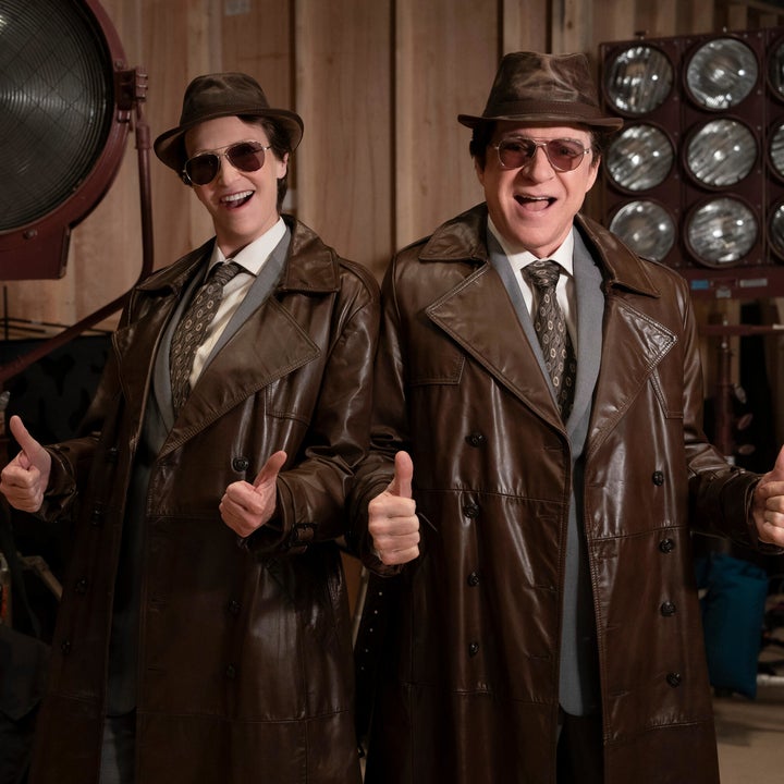 Two individuals wearing sunglasses, brown trench coats, and matching hats give thumbs-up gestures on a set resembling a film studio