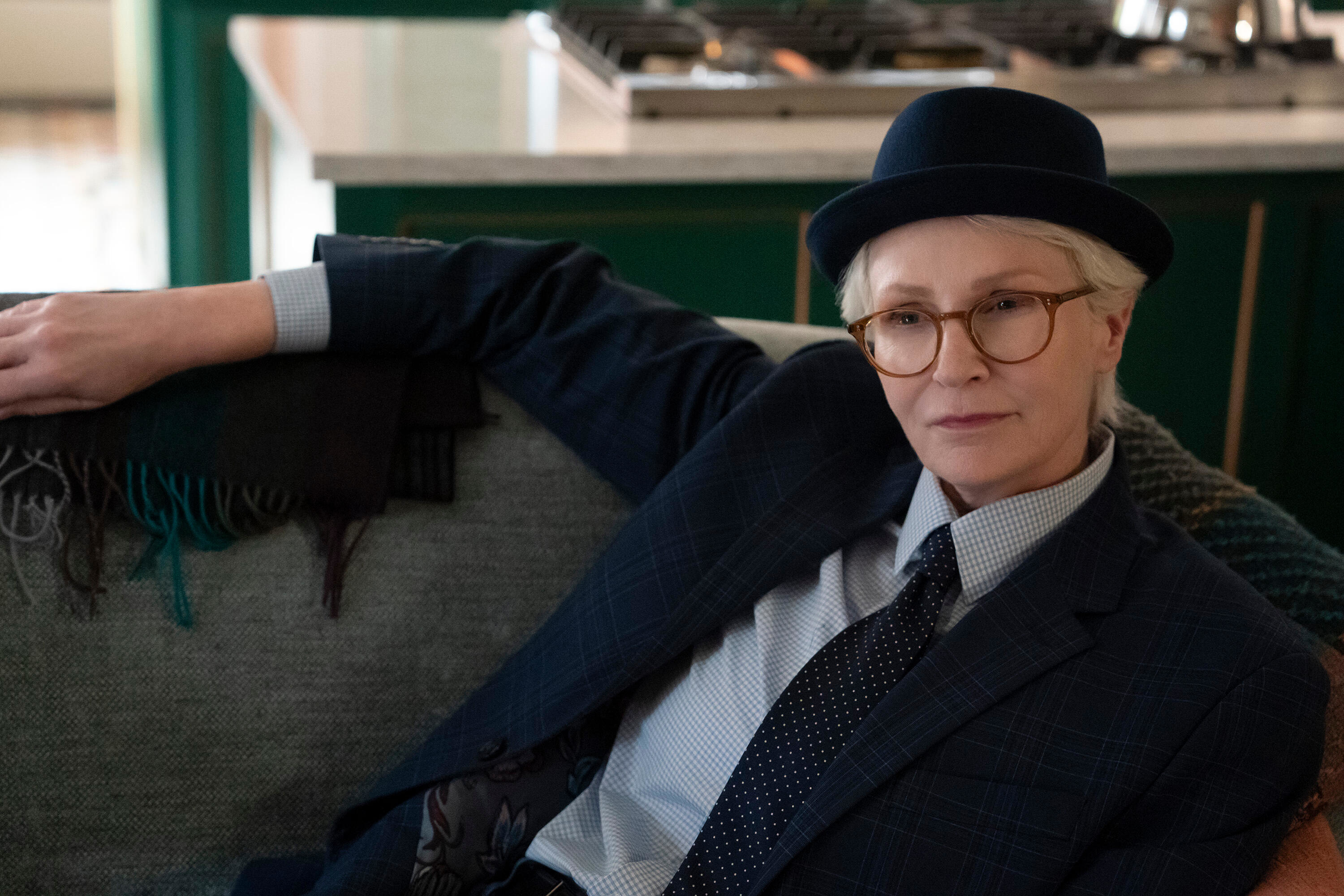Jane Lynch, dressed in a suit and hat, lounges on a couch in a well-lit living area