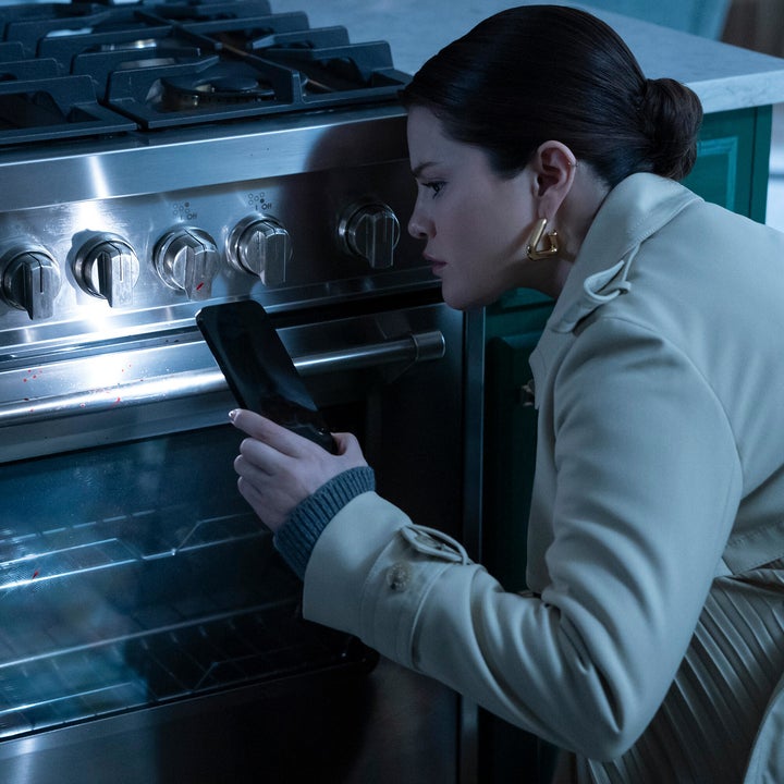 A focused Selena Gomez examines a modern stainless steel oven with her smartphone, dressed in an elegant trench coat