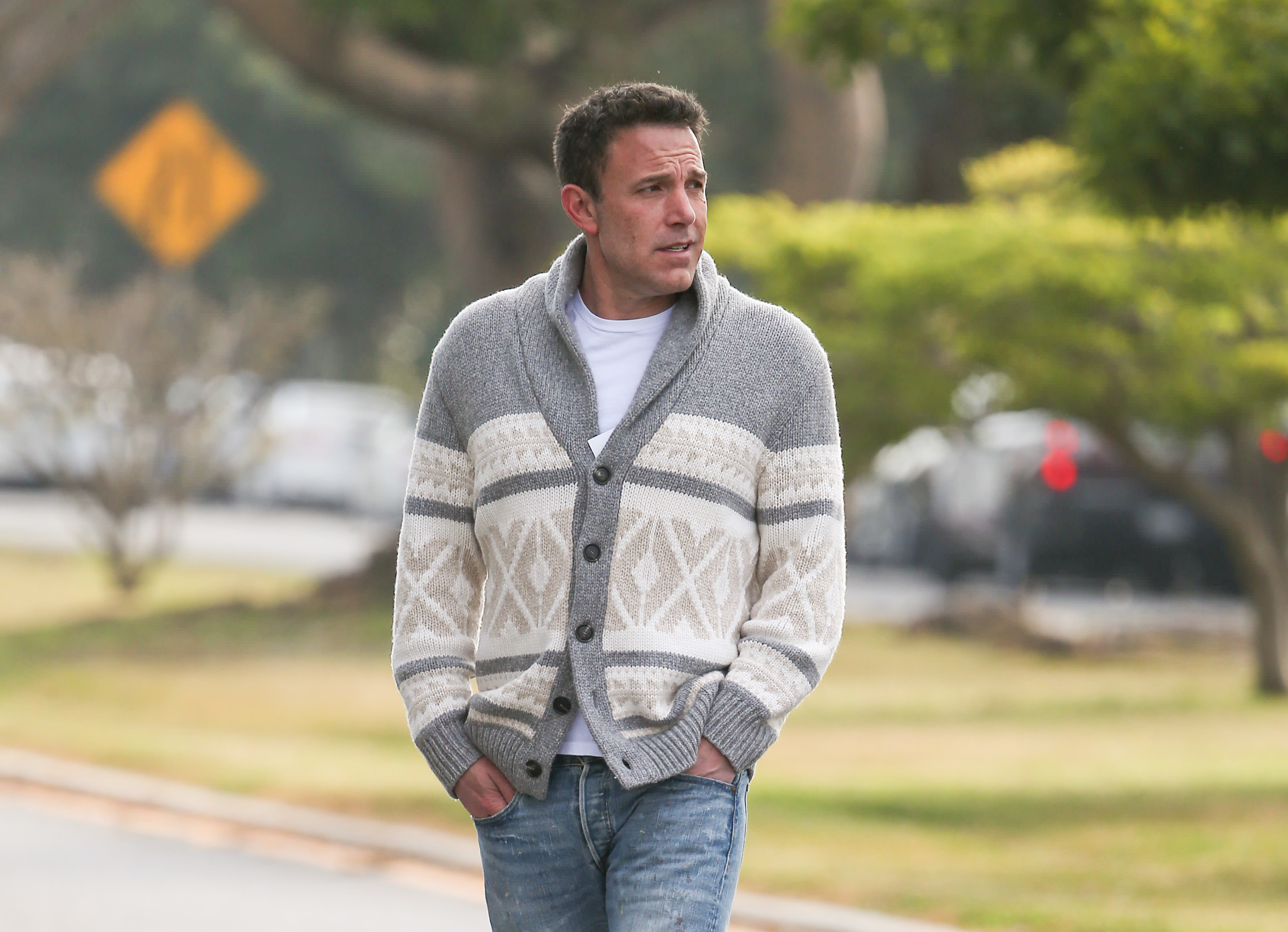 Ben Affleck walking outdoors wearing a casual patterned cardigan, white t-shirt, and jeans with hands in pockets