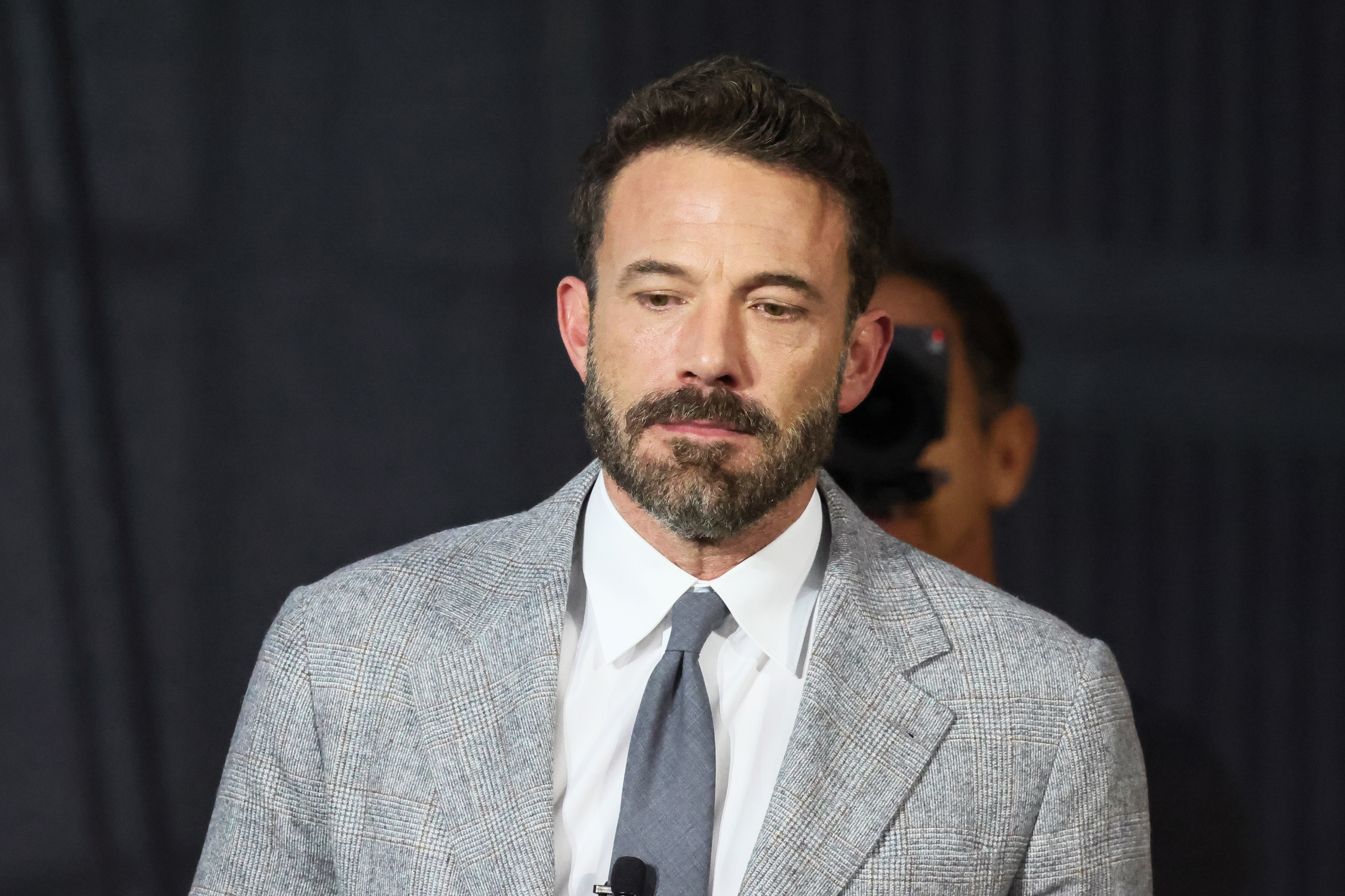 Ben Affleck is pictured wearing a gray suit with a white shirt and a dark tie, standing against a plain background