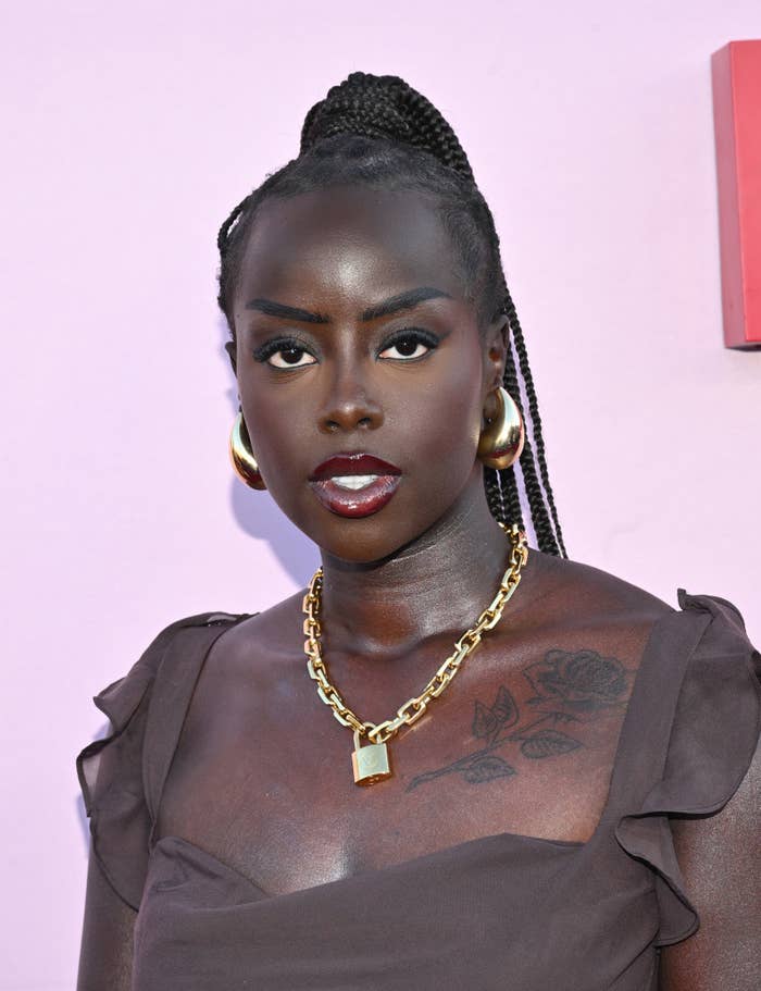 Golloria with braided hair, gold hoop earrings, and a gold necklace with a pendant, wearing a sleeveless top. There is a floral tattoo on her left shoulder.