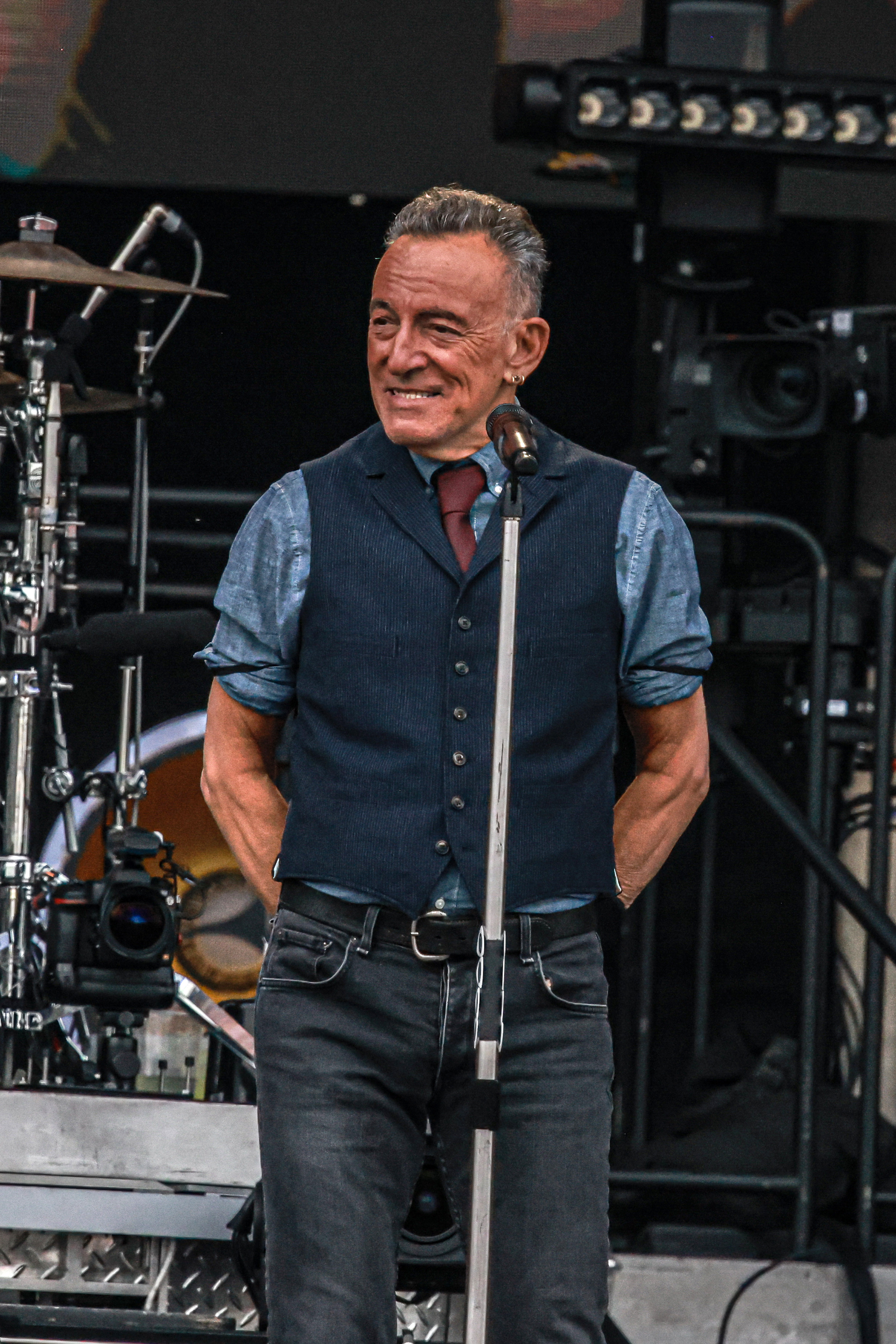Bruce Springsteen stands on stage in front of a microphone, wearing a vest, shirt, tie, and jeans, smiling during a performance