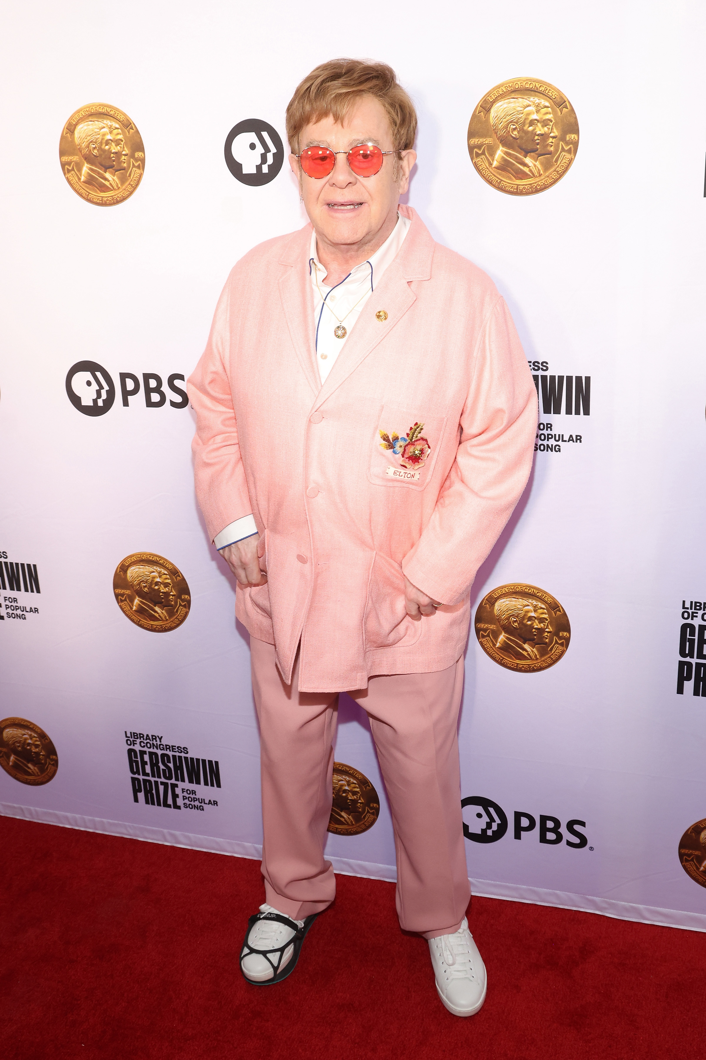 Elton John on the red carpet in a loose pink suit with embroidered details, white shirt, and pink trousers, wearing red-tinted glasses and black and white sneakers
