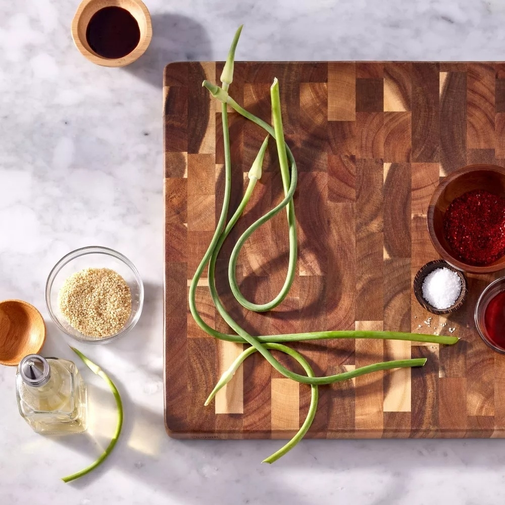 Chopping board with garlic scapes, sesame seeds in a glass bowl, soy sauce, chili powder, salt, and oil arranged around it