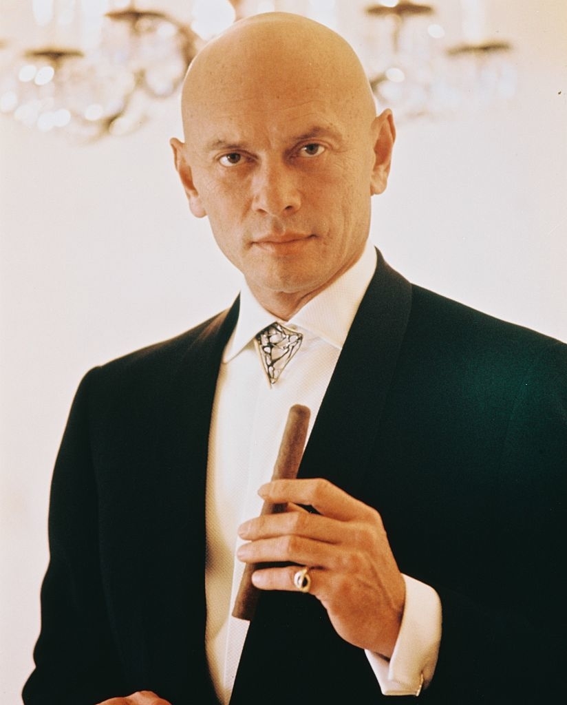 A bald man in formal attire holds a cigar while looking straight at the camera. An elegant chandelier is visible in the background