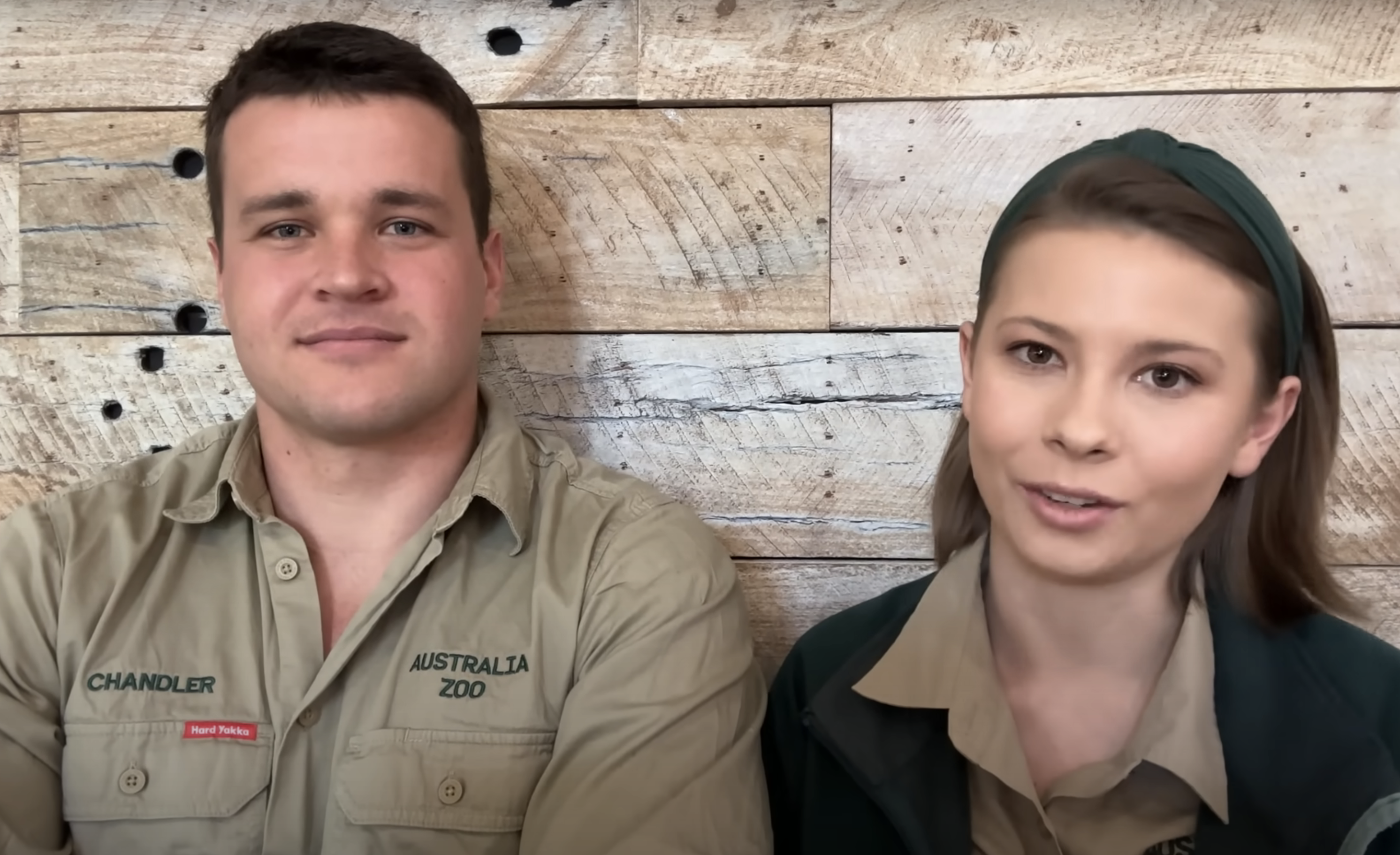 Chandler Powell and Bindi Irwin wear Australia Zoo uniforms and smile while posing against a wooden backdrop