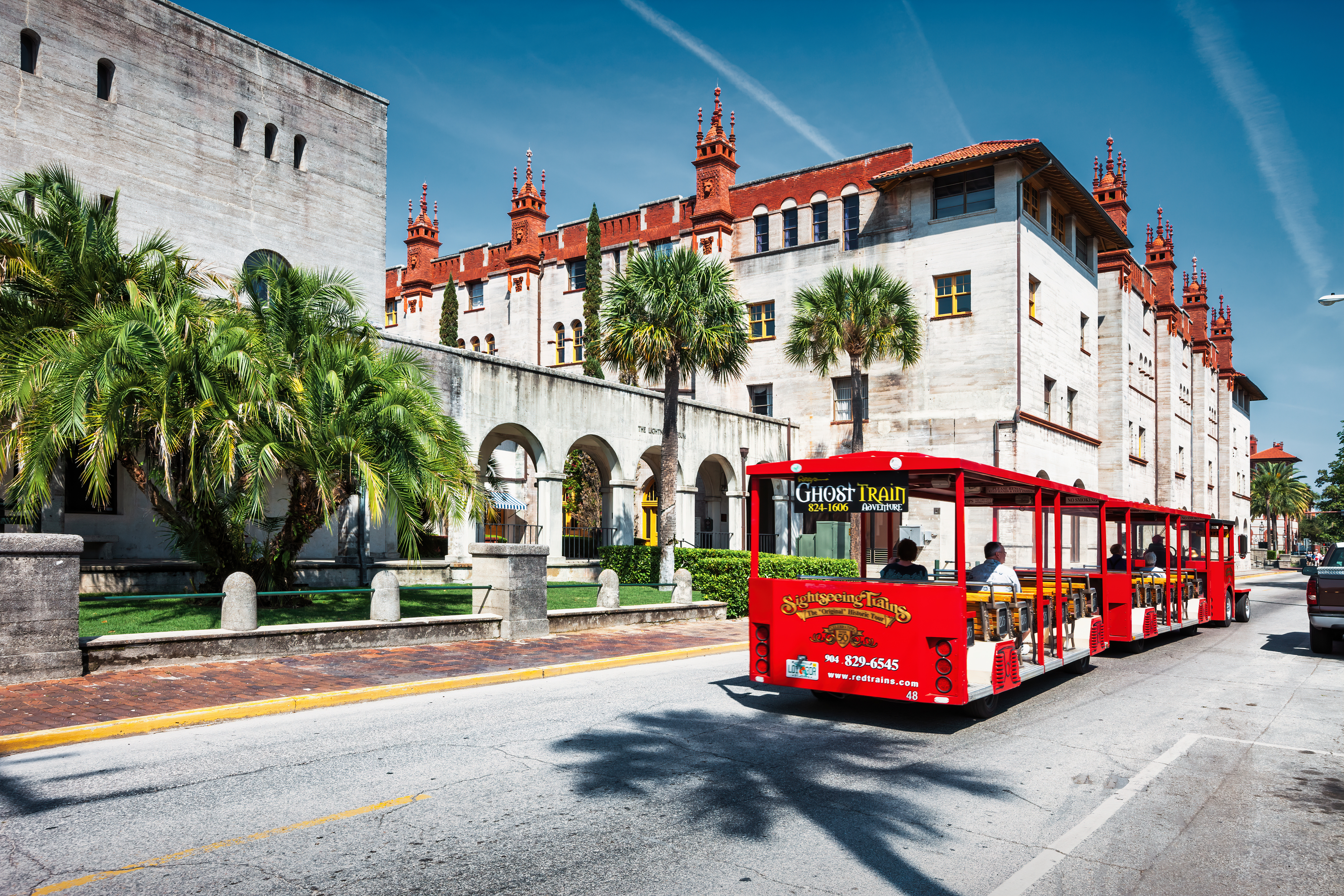 A red trolley rides past a historic, castle-like building with palm trees lining the street in St. Augustine, Florida