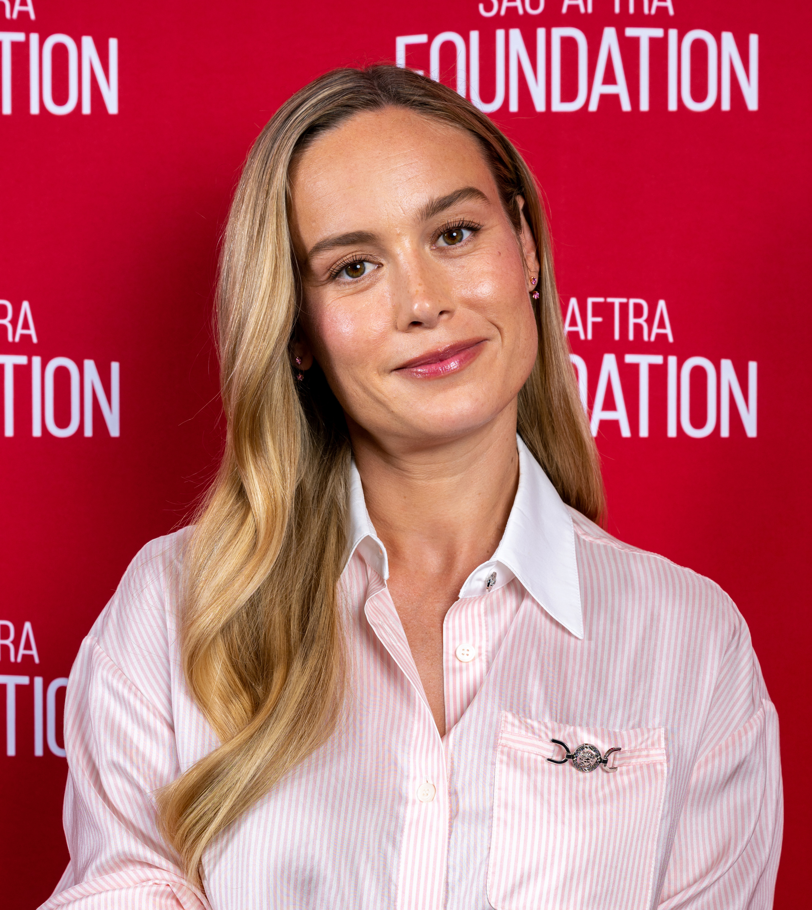 Brie Larson at a SAG-AFTRA Foundation event, wearing a white blouse with subtle pinstripes. She has long, wavy hair styled down. The background reads &quot;SAG-AFTRA Foundation&quot;