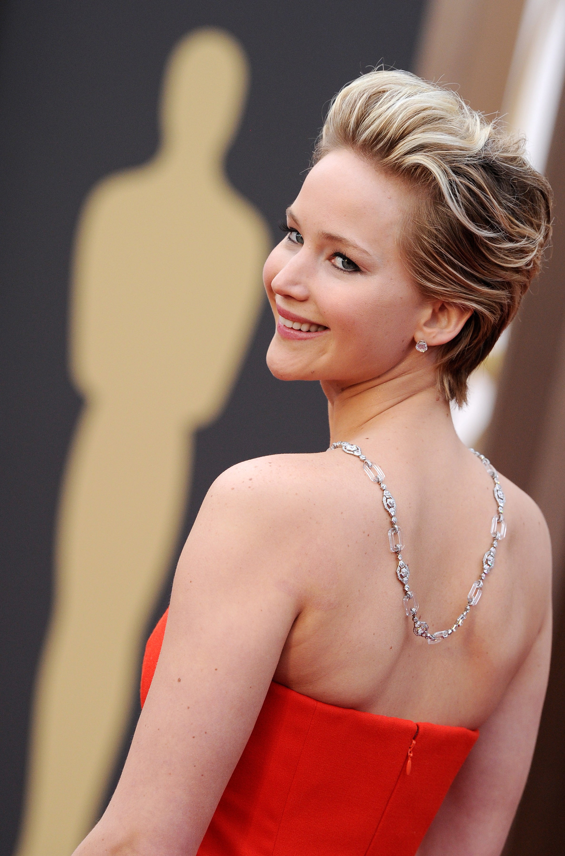 Jennifer Lawrence poses on a red carpet wearing a strapless gown, smiling over her shoulder. An Oscar statue is blurred in the background