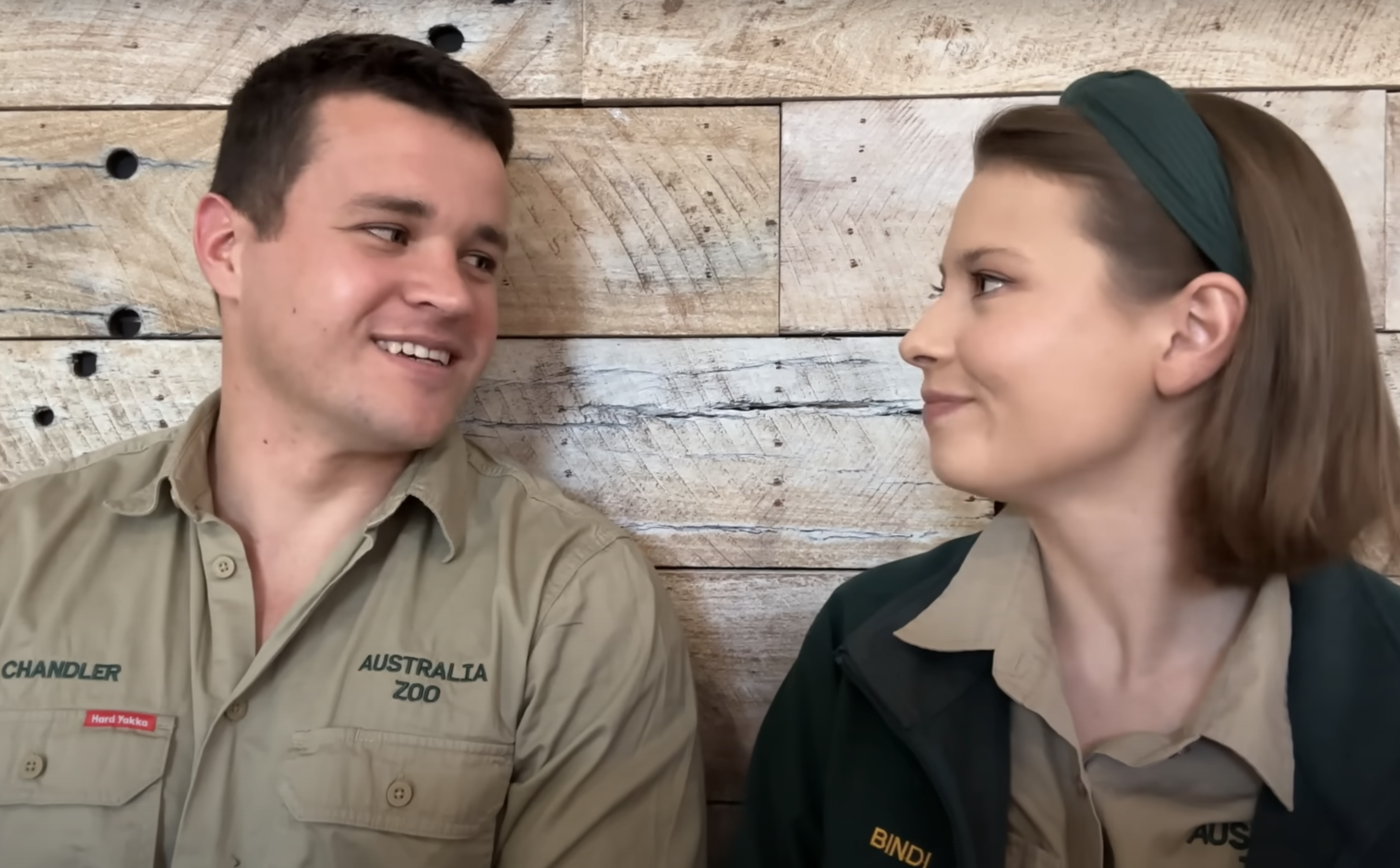Chandler Powell and Bindi Irwin, both in uniform from Australia Zoo, smile at each other in front of a wooden backdrop
