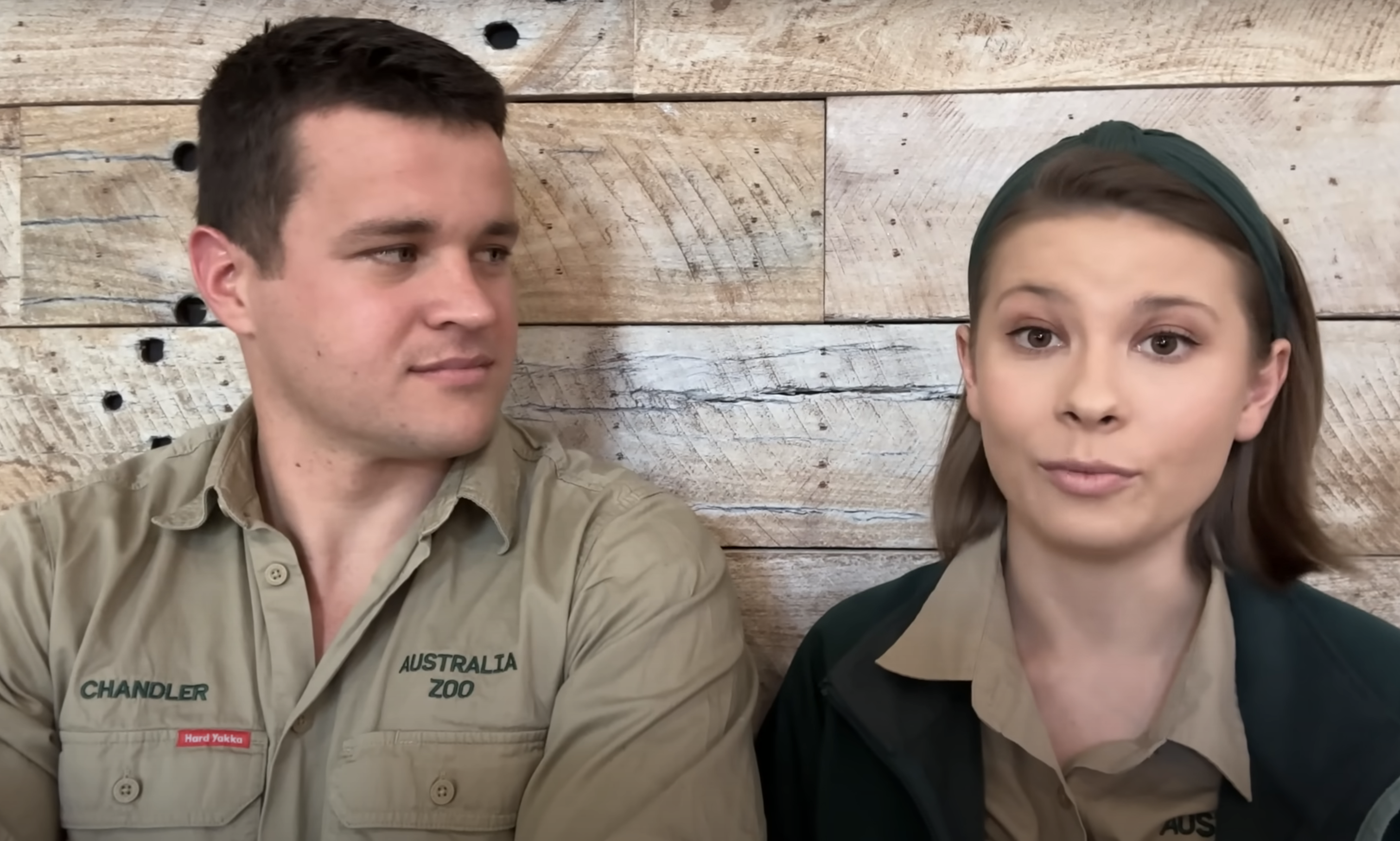 Chandler Powell and Bindi Irwin in Australia Zoo uniforms, sitting against a wooden backdrop, looking at each other