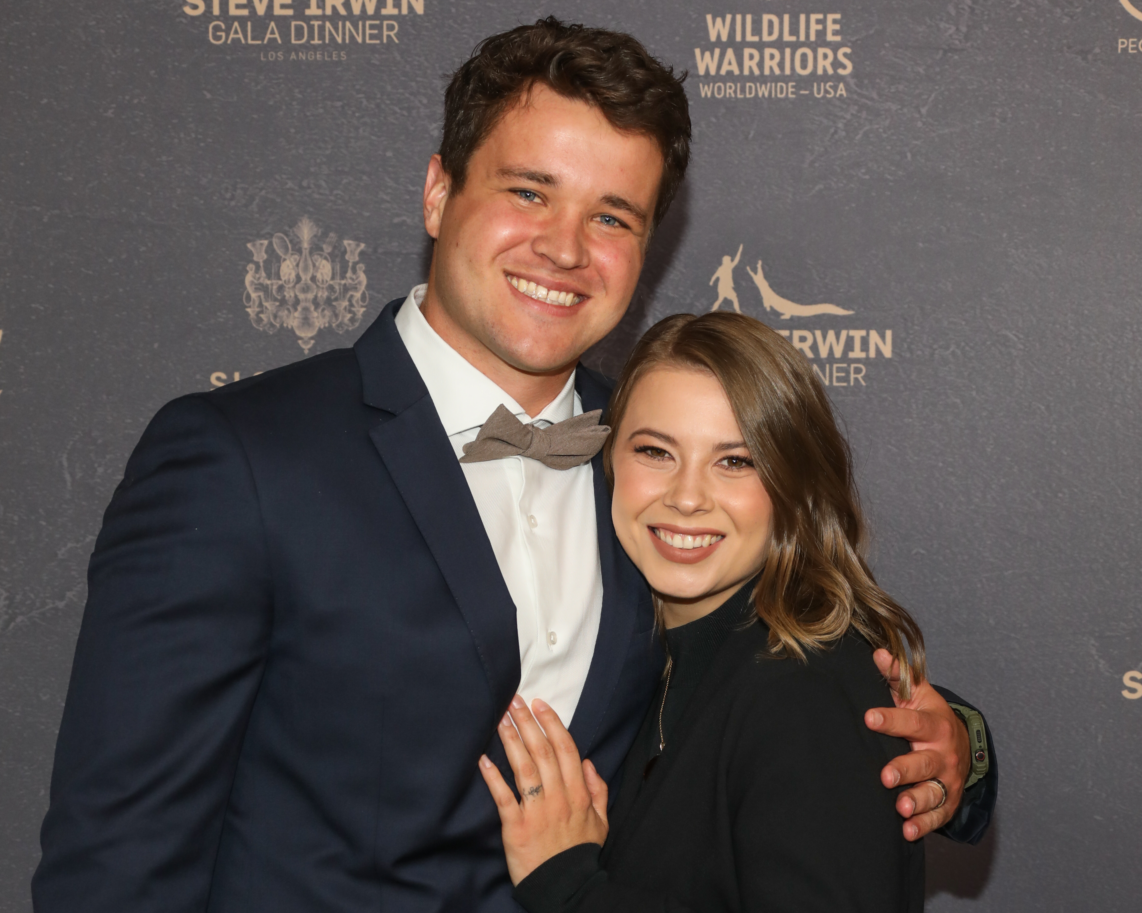 Chandler Powell and Bindi Irwin smile and pose together at the Steve Irwin Gala Dinner. Chandler wears a suit with a bow tie, and Bindi wears a dark outfit
