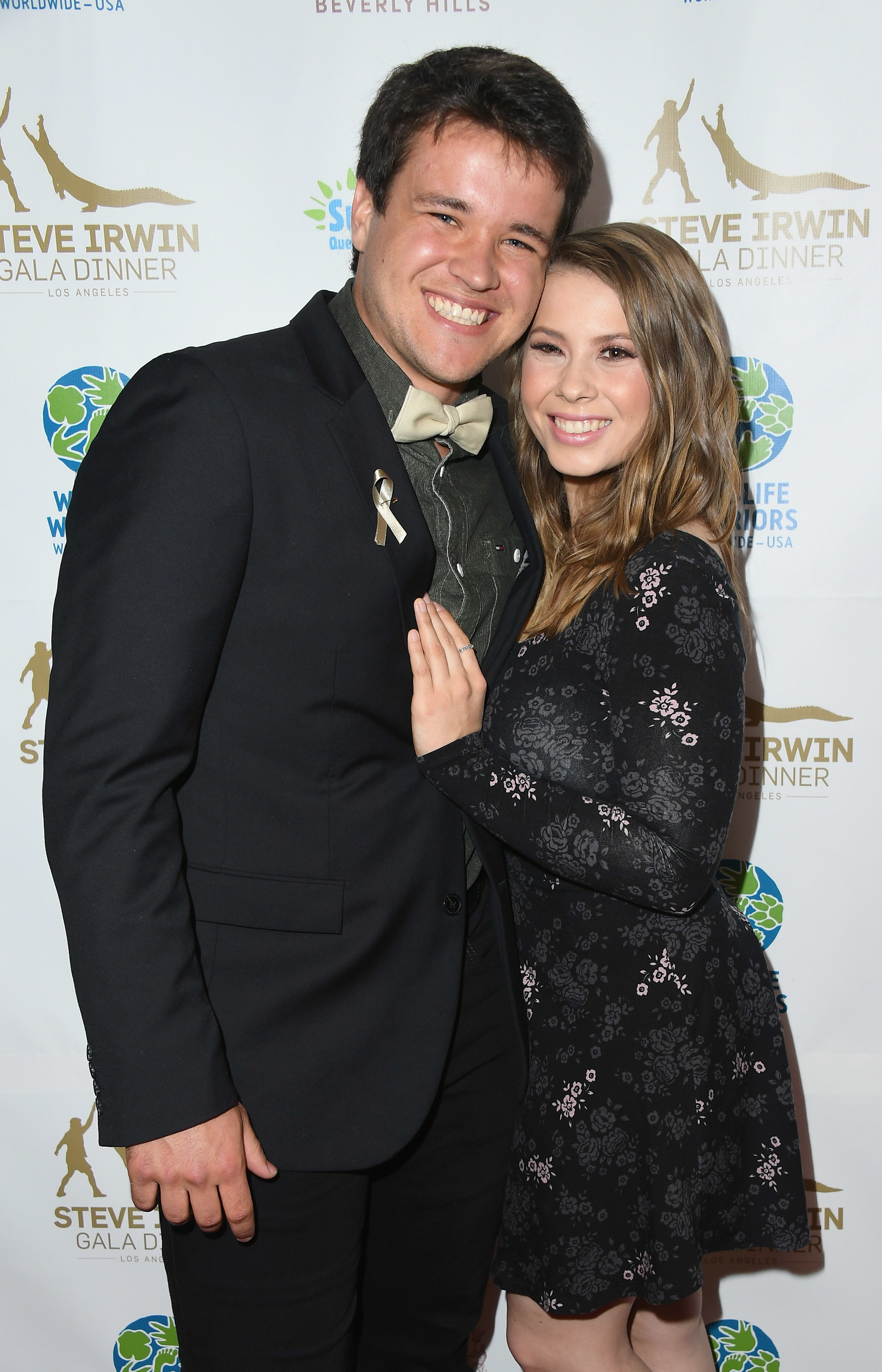Chandler Powell and Bindi Irwin smiling and posing together on the red carpet at the Steve Irwin Gala Dinner. Chandler is in a suit, and Bindi wears a floral dress