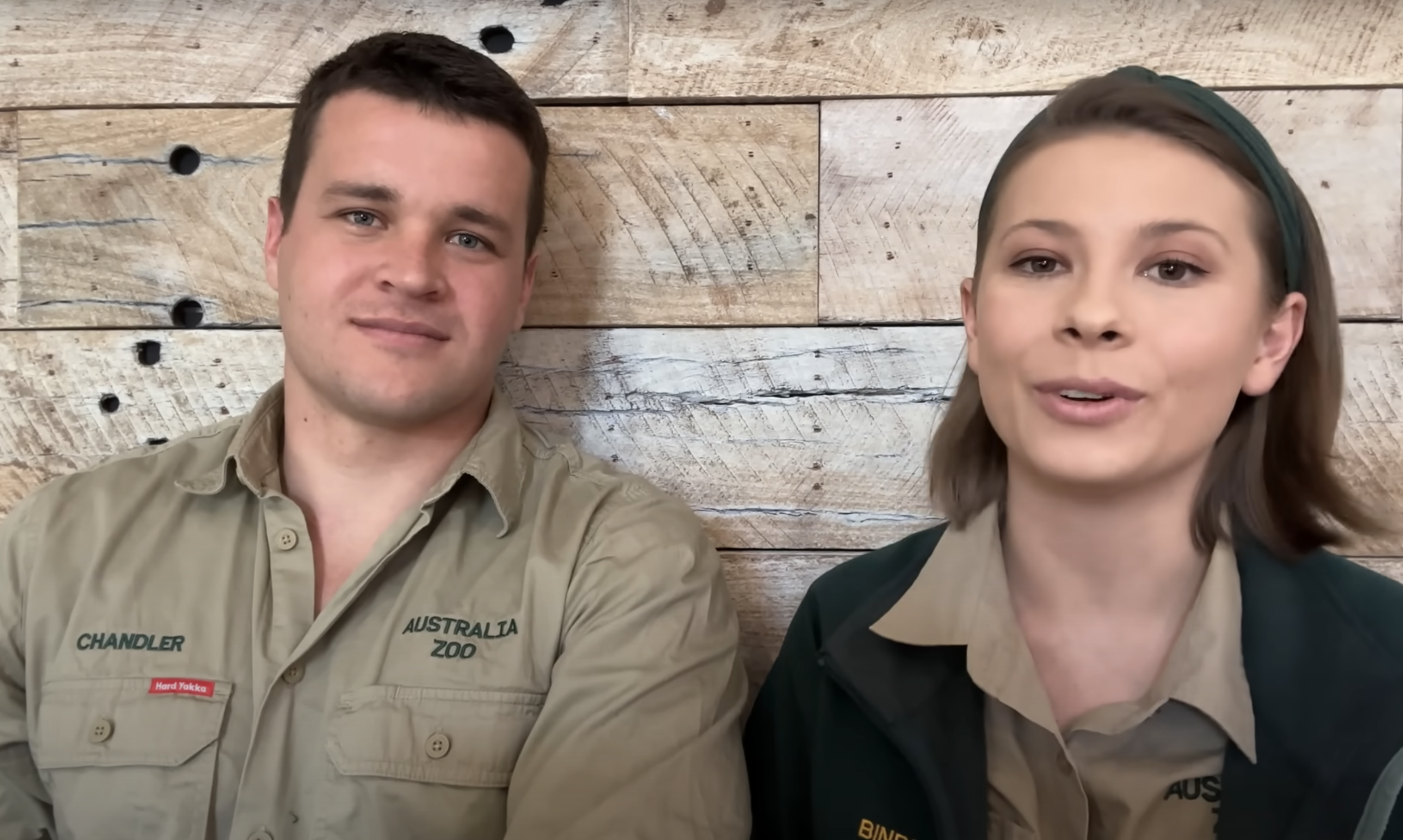 Chandler Powell and Bindi Irwin wear Australian Zoo uniforms, sitting in front of a wood-paneled wall