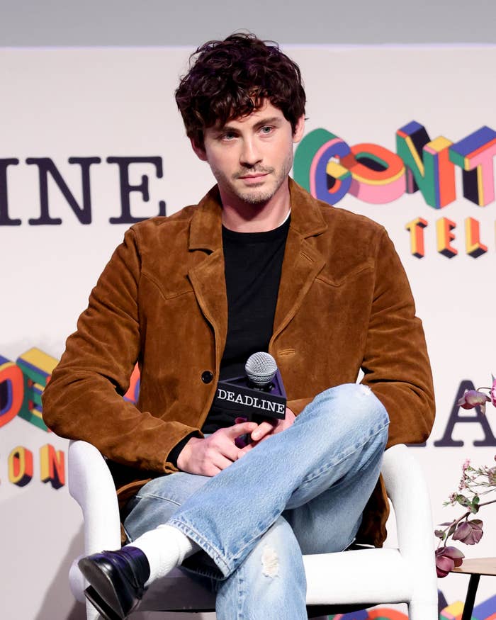 Logan Lerman at a Deadline event, wearing a suede jacket, dark shirt, and light blue jeans, holding a microphone