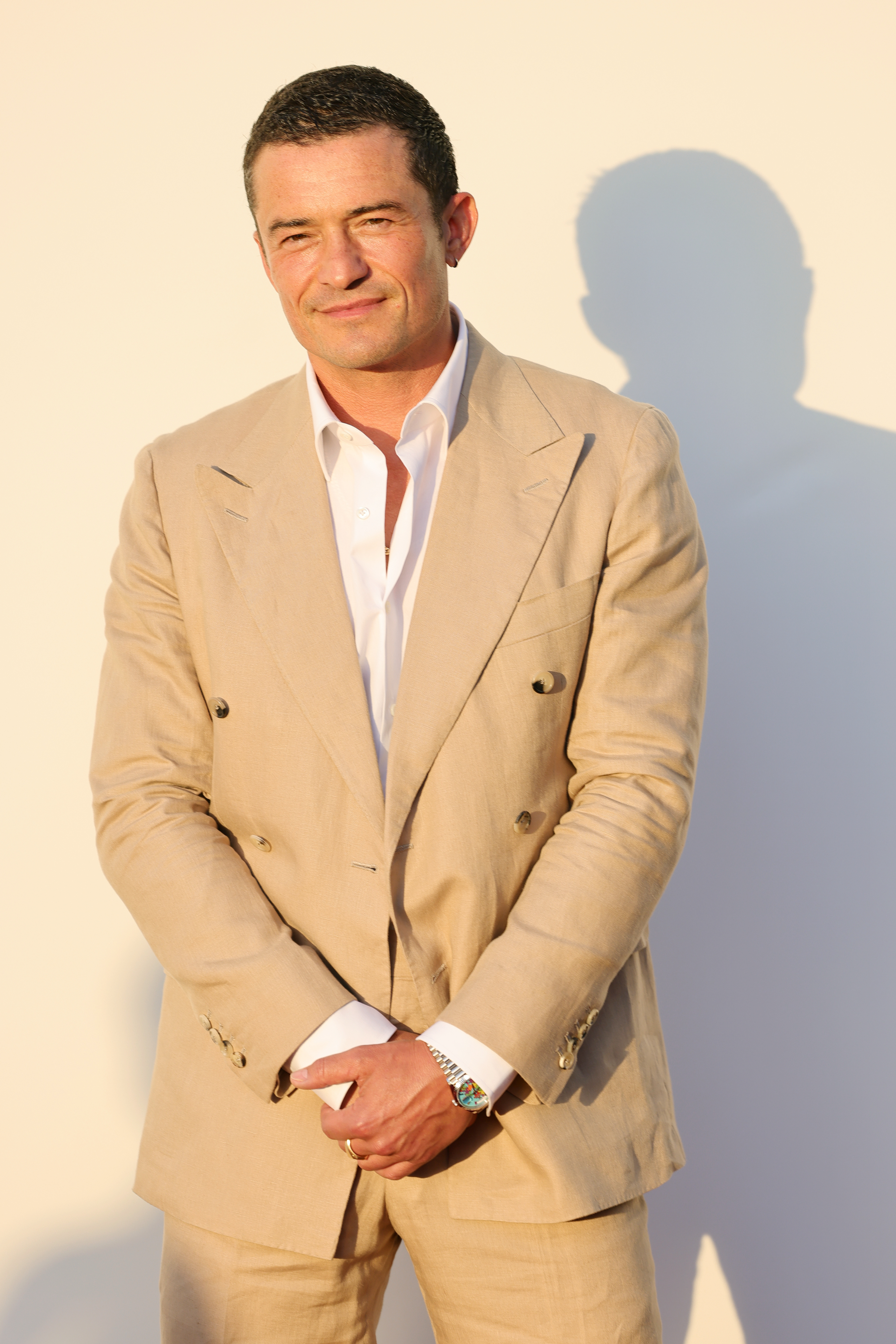 Orlando Bloom in a tan double-breasted suit with a white shirt, standing against a plain background
