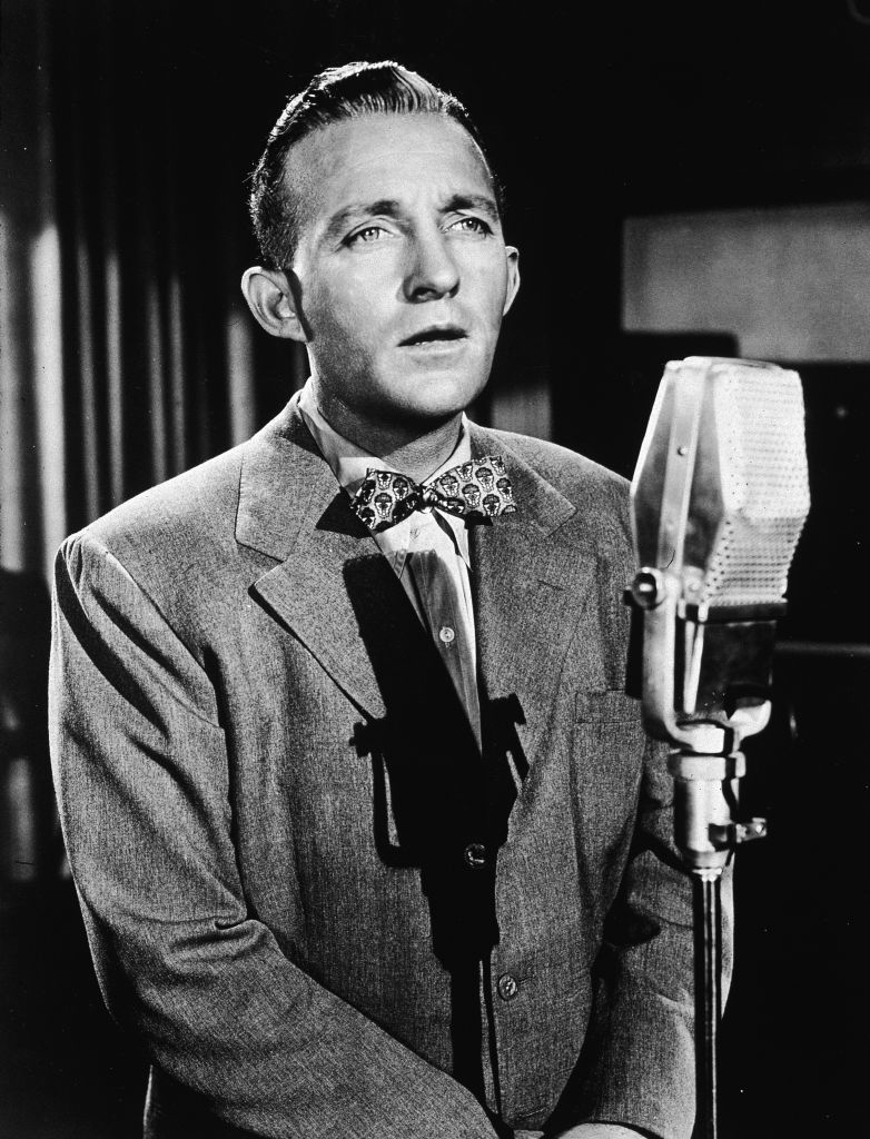 A man stands in front of an old-fashioned microphone, wearing a suit and a bow tie, singing or speaking