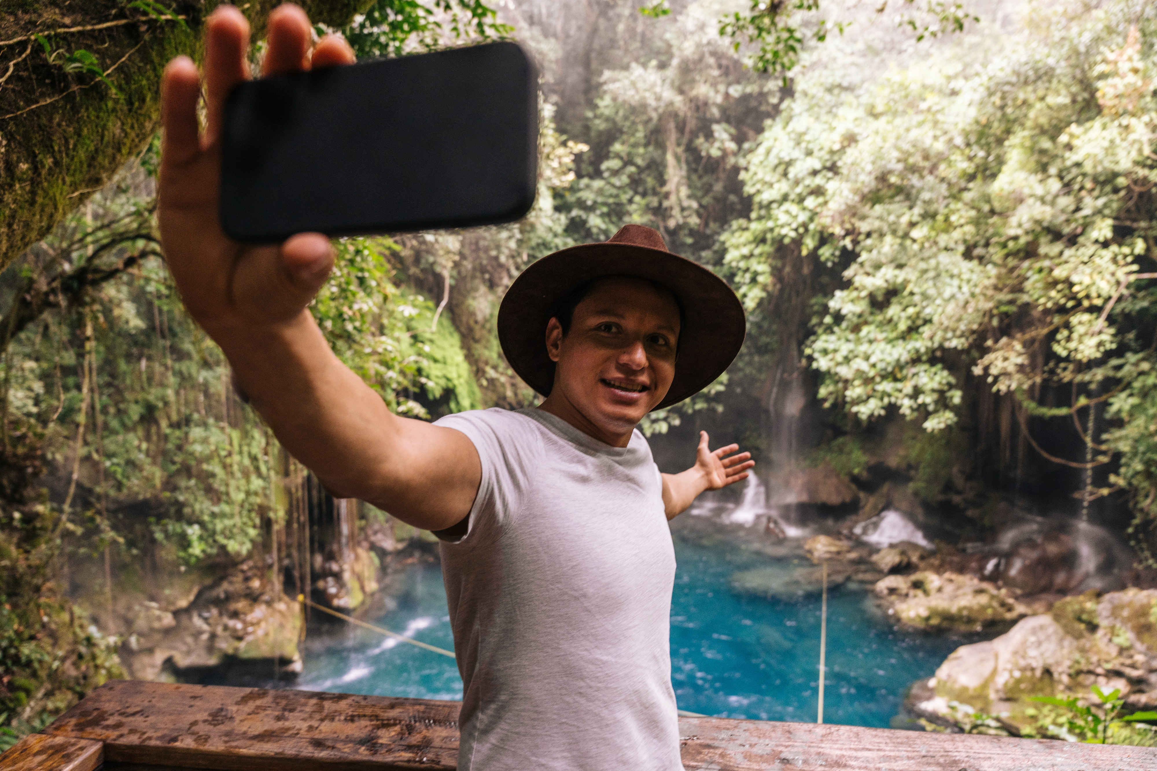 Man in a t-shirt and hat takes a selfie in front of a waterfall and lush greenery in Sonora, Mexico