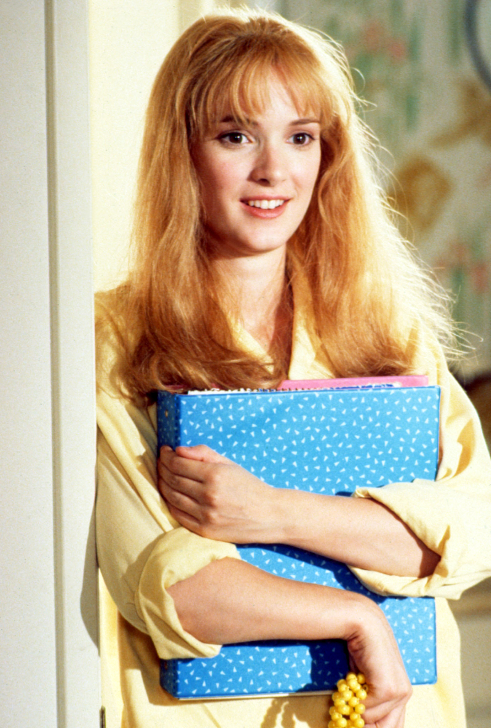 Winona Ryder wearing a yellow top, holding a blue binder while leaning against a wall, smiling in "Edward Scissorhands"