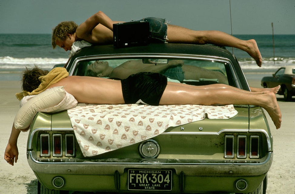 Three people relax on the trunk and roof of a car with a Michigan license plate at the beach. Two are lying on their stomachs, and one is standing, leaning over the roof