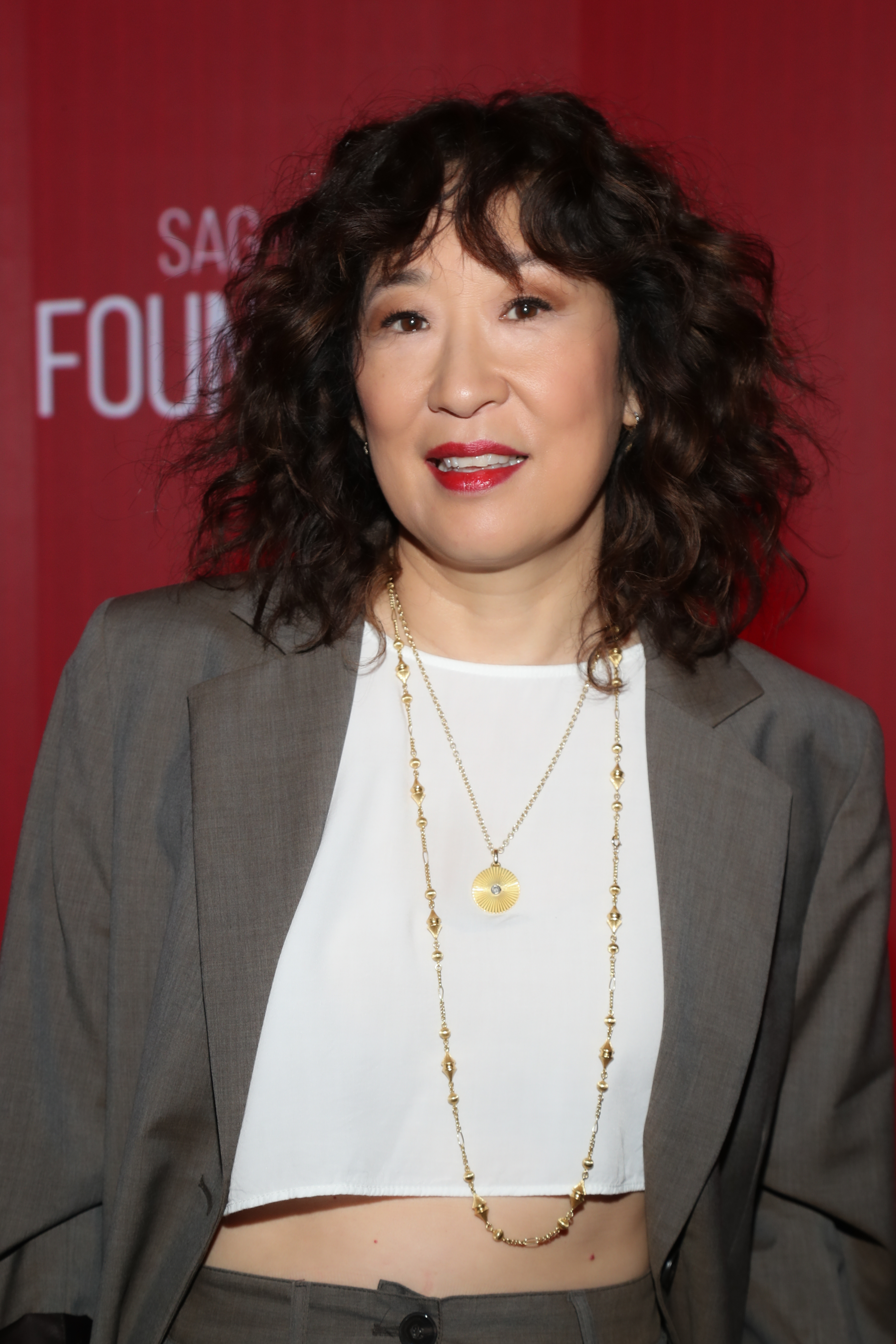 Sandra Oh in a cropped white top with a long necklace, neutral blazer, and matching trousers poses in front of a SAG-AFTRA Foundation backdrop