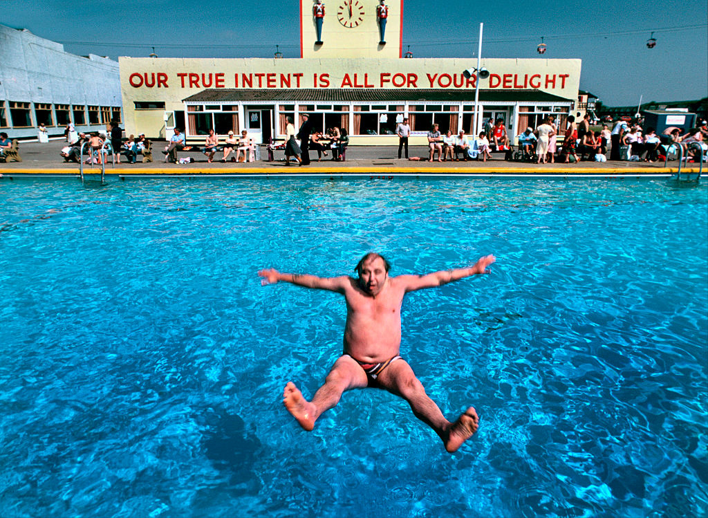 An elderly man jumps backwards into a pool in Skegness, UK, with a crowd seated in the background near a building, reading &quot;Our true intent is all for your delight&quot;