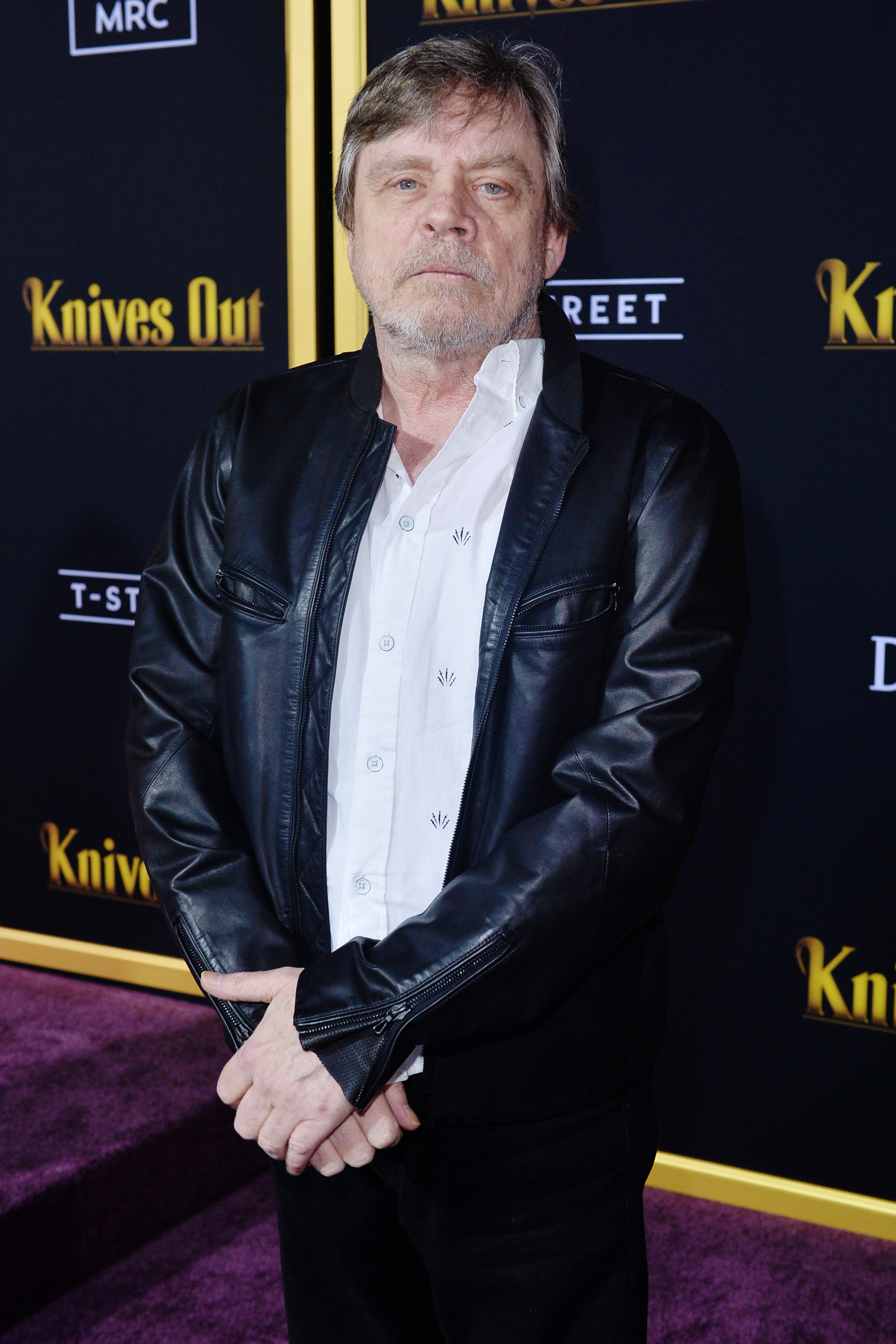 Mark Hamill on the red carpet for "Knives Out" premiere, wearing a black leather jacket over a white button-up shirt