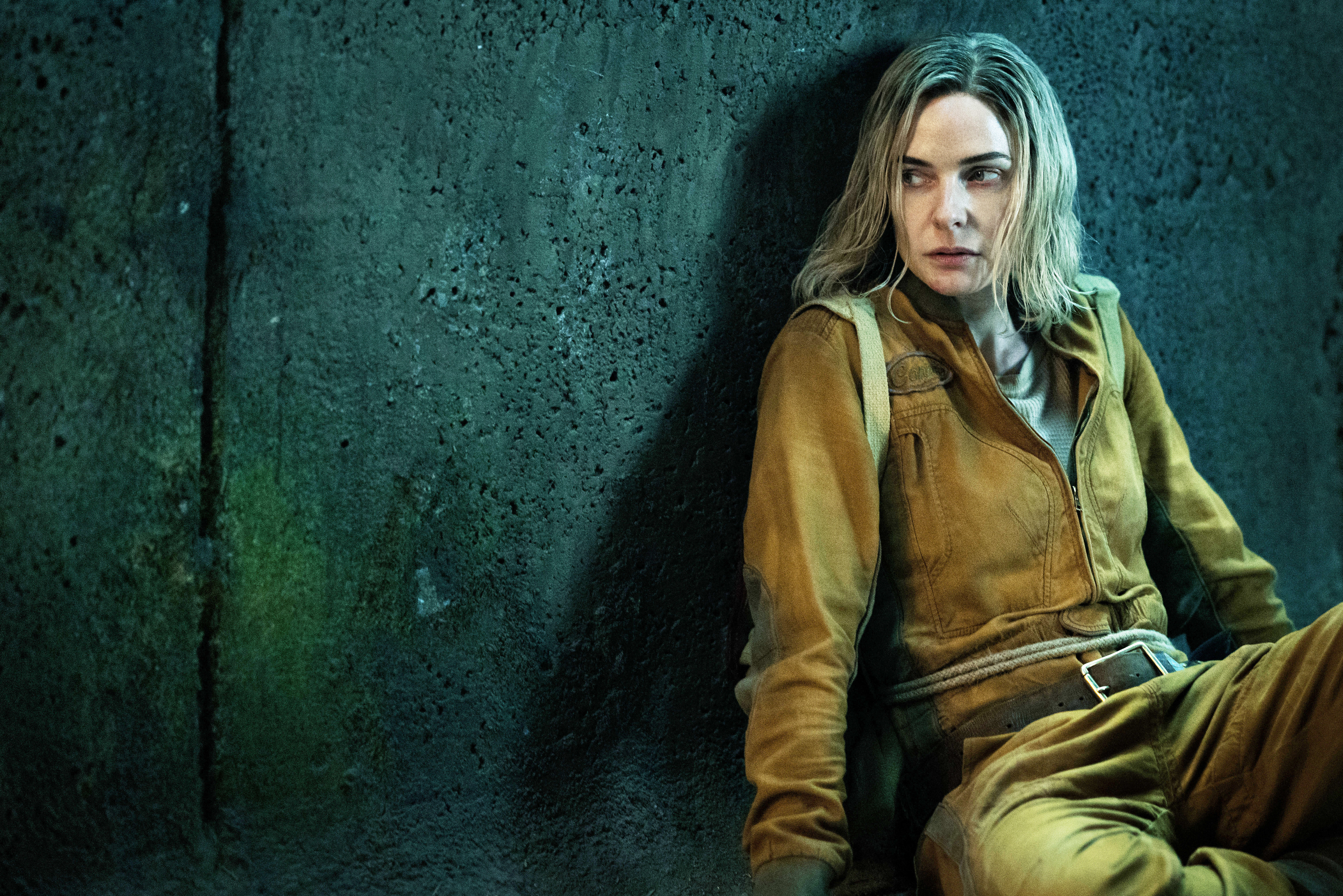Rebecca Ferguson sits against a rough concrete wall, wearing utilitarian clothing, with a worn-out expression, likely depicting a scene from a film or TV show