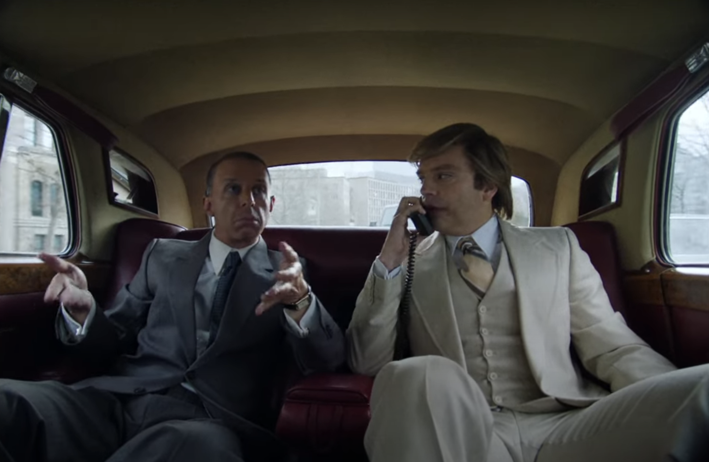 Jeremy Strong and Sebastian Stan sit in the back of a car, Bob speaking on an old-fashioned car phone, both dressed in suits