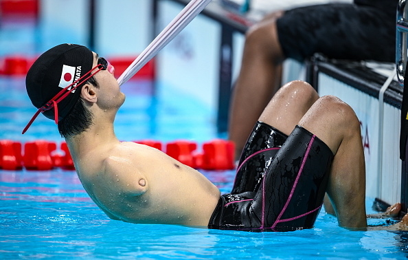 Male swimmer in swim cap and goggles prepares for a race, gripping lane marker with teeth, legs bent at poolside