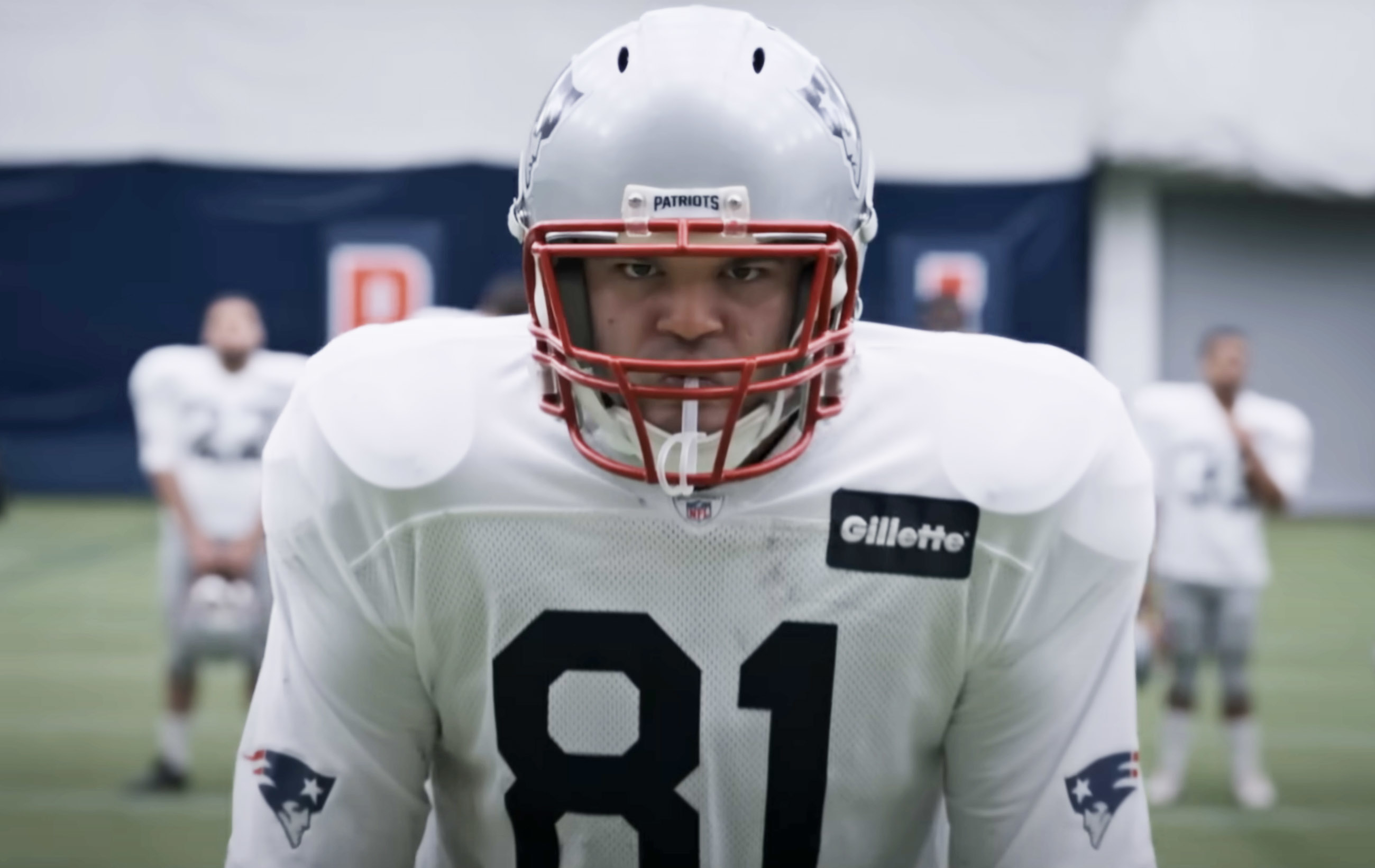 Football player Aaron Hernandez in a New England Patriots uniform with number 81 and a helmet is on the field, looking intensely into the camera in a scene from "American Sports Story"