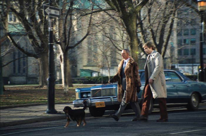 Two men walk a dog on a city street. One man wears a long fur coat, and the other wears a beige trench coat. A vintage car is parked behind them