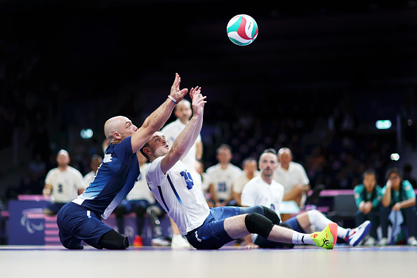 Two male sitting volleyball players in action on the court, attempting to reach the ball mid-air. The foreground player is in a blue uniform, and the background player is in a white uniform