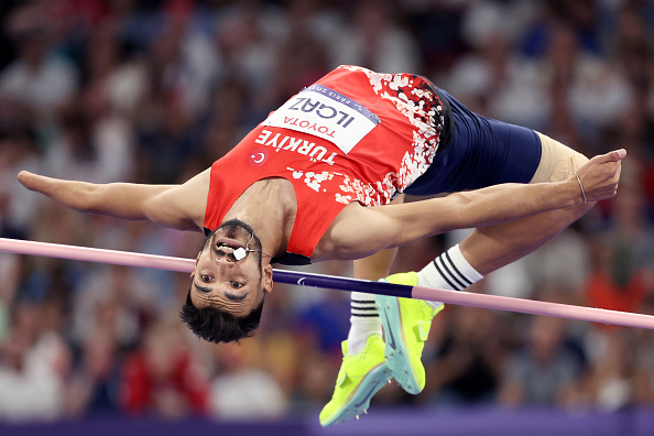 Husnu Can Ozbek competes in a high jump event, wearing athletic gear and yellow shoes, clearing the bar with a Fosbury Flop technique at a competition