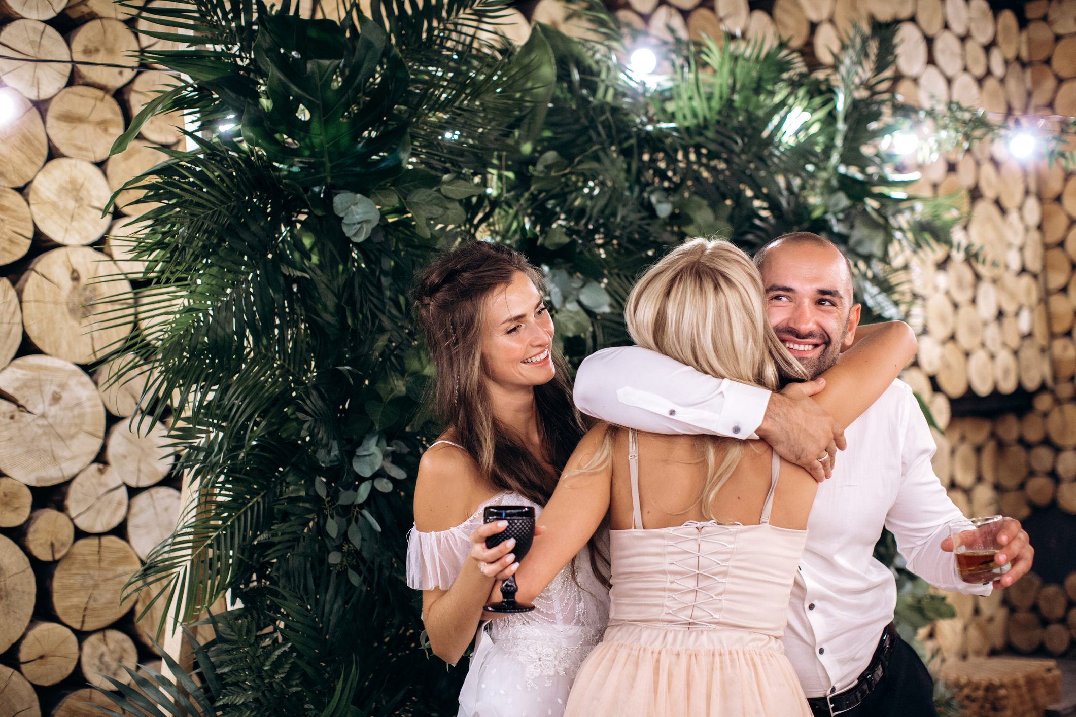 Two women and a man in formal attire embrace joyfully at a wedding celebration; the setting features lush greenery and wooden logs