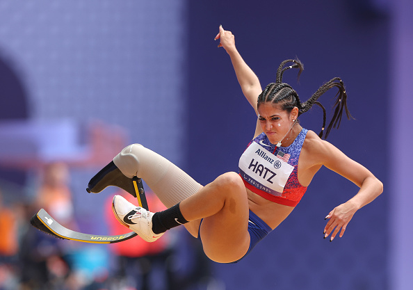 Athlete Scout Bassett, wearing a blue and red athletic outfit, competes in a long jump event using a prosthetic leg