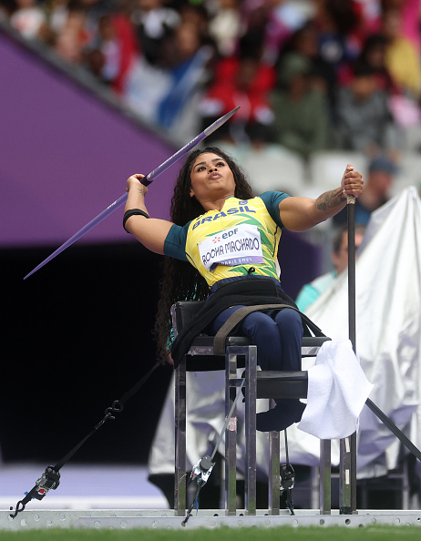 Rocia Machado competes in a javelin throw event. She is captured mid-throw on a chair, wearing athletic attire with her name displayed on her chest