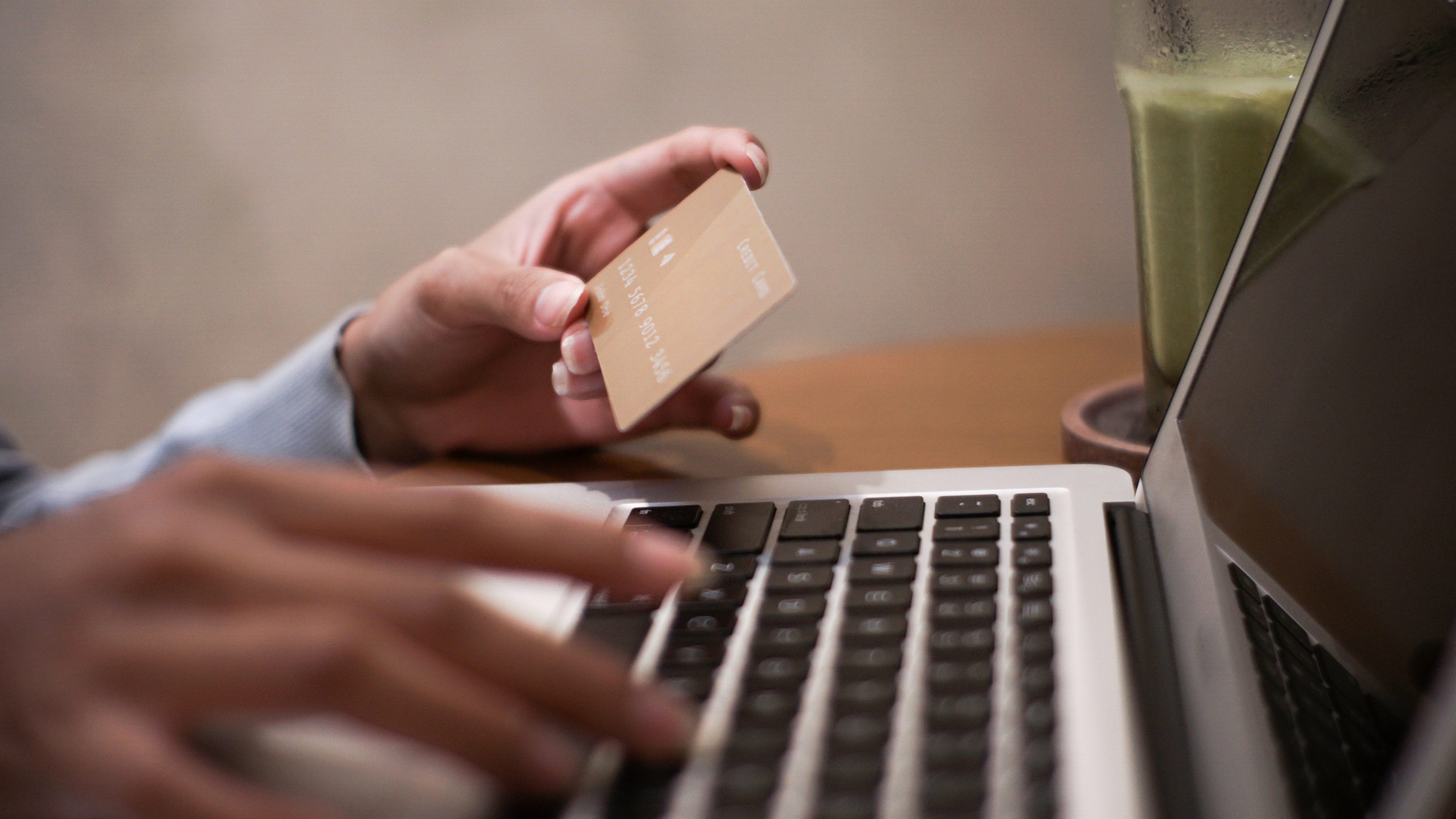Person typing on a laptop with one hand while holding a credit card in the other, possibly making an online purchase. A glass is nearby