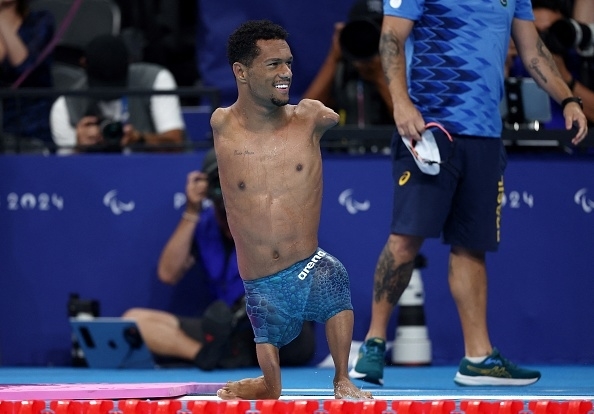 Swimmer with limb differences stands by the pool in competition attire at a Para swimming event, with other participants and photographers in the background