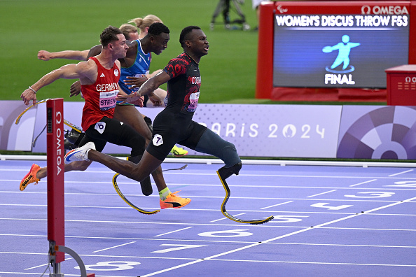 Several runners, including Johannes Floors, compete in a track event at the Paris 2024 Paralympic Games. A 'Women's Discus Throw F53 Final' sign is visible in the background