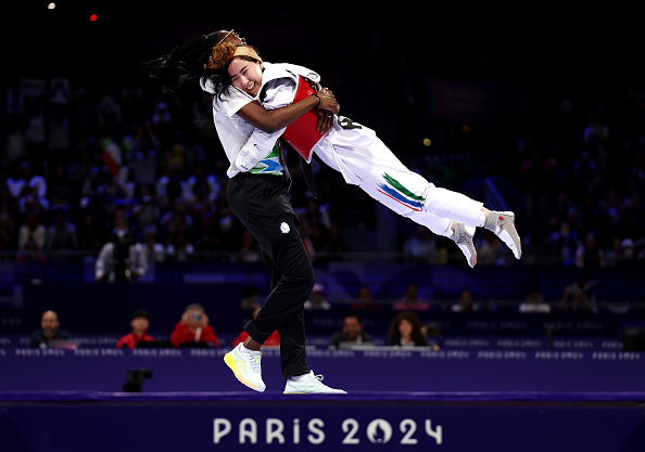 A joyful athlete lifts another in celebration after a match at the Paris 2024 event. Both are in white athletic gear. The background shows an audience and the Paris 2024 sign