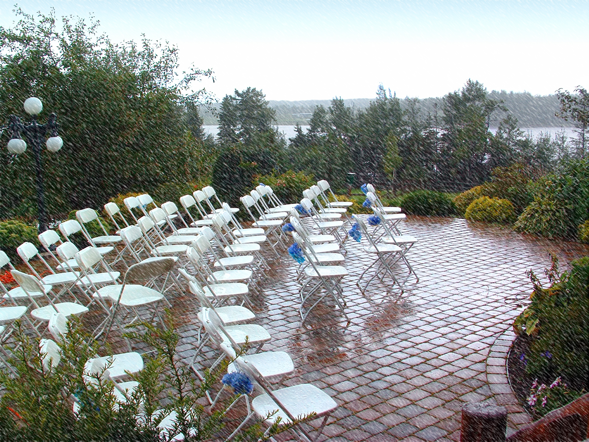 Outdoor wedding setup with rows of empty white chairs arranged on a brick patio, with a scenic lake and trees in the background, captured during rainfall