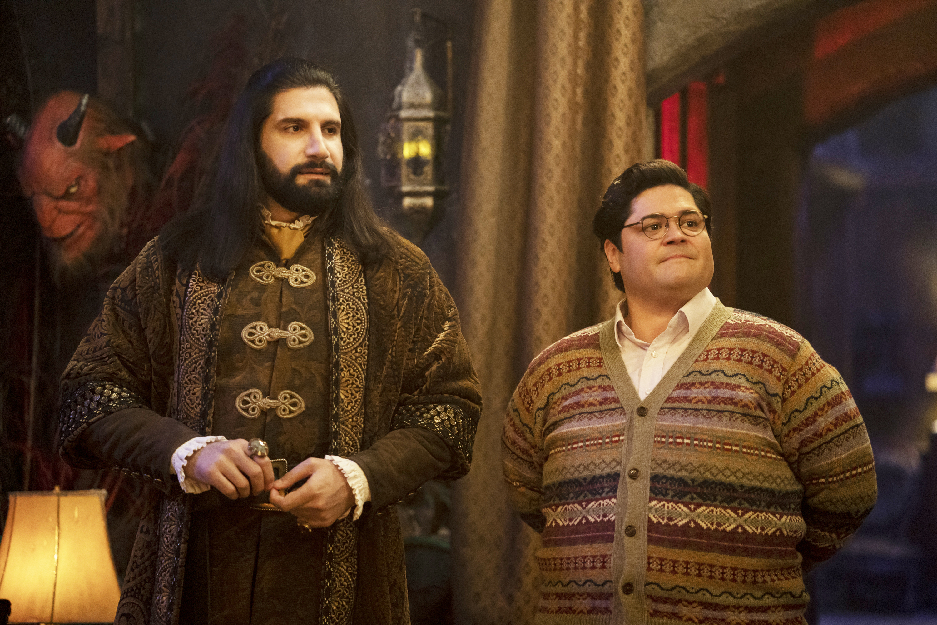Kayvan Novak in a medieval-style outfit stands beside Harvey Guillén, who is wearing a patterned sweater, on the set of "What We Do in the Shadows."