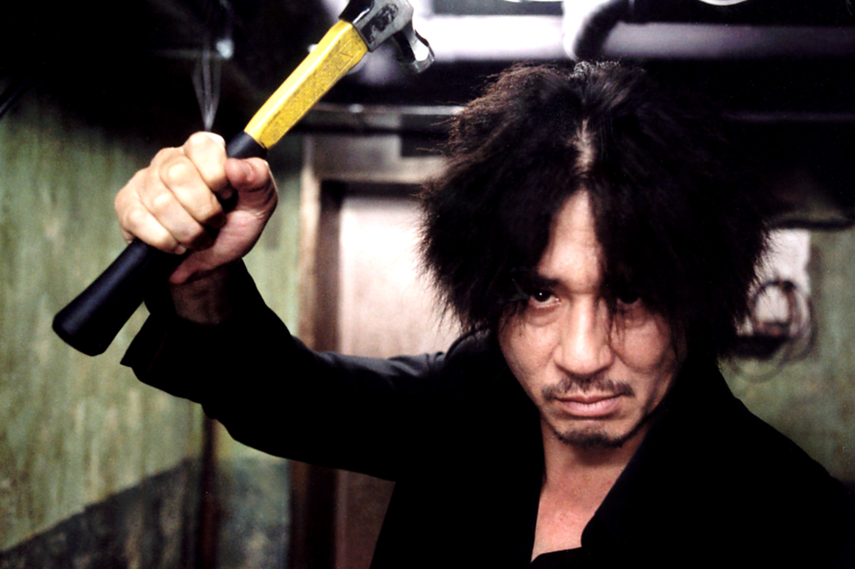 A man with disheveled hair, intense expression, and dark clothing holds a hammer above his head in a threatening manner, in a dimly lit hallway
