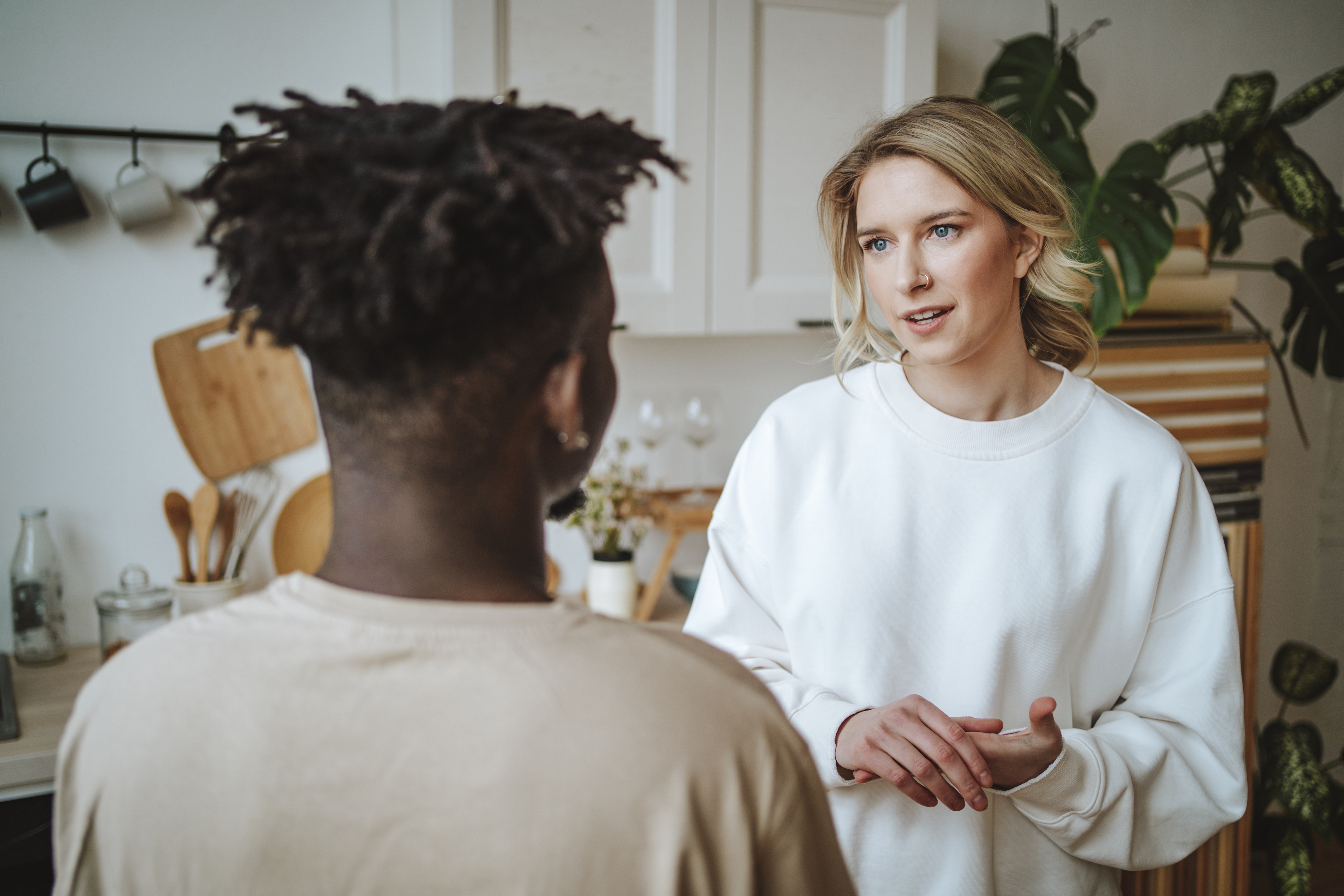 A man and a woman are having a serious conversation in a kitchen. The woman is wearing a casual long-sleeve shirt and the man has his back to the camera