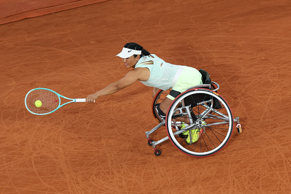 A wheelchair tennis player in action on a clay court, reaching out to hit a tennis ball with their racket, displaying athleticism and focus
