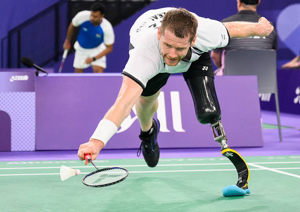 David Phillips, a para-badminton player with a prosthetic leg, lunges to hit a shuttlecock during a match. An opponent and a referee are visible in the background